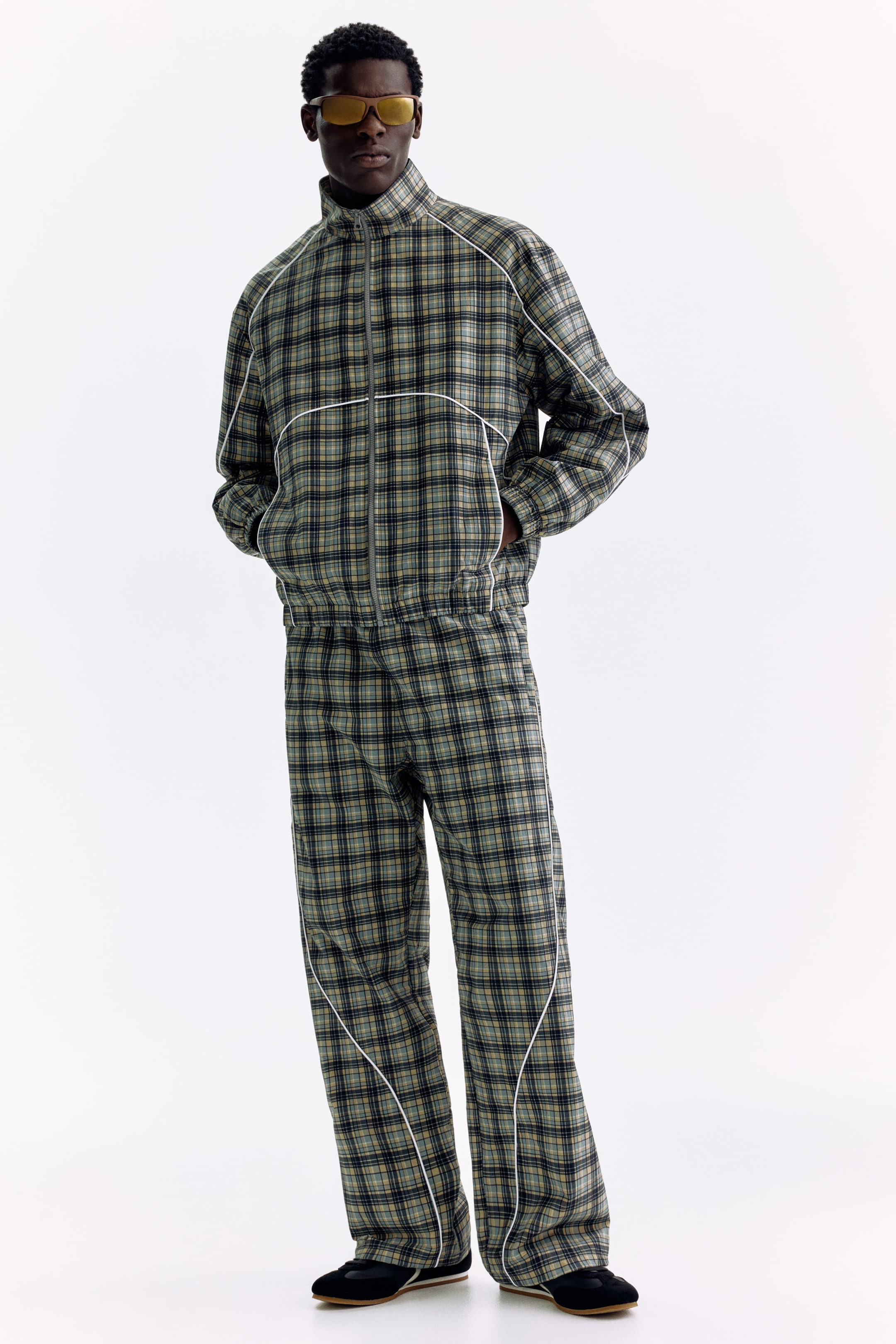 Ampliar la imagen: Un conjunto deportivo de cuadros en verde, azul y beige está siendo exhibido, compuesto por una chaqueta con cremallera completa, cuello alto y dobladillo elástico, y pantalones de pierna ancha a juego. Un ribete curvado blanco detalla la chaqueta a lo largo del pecho y los pantalones en la parte delantera de las piernas.