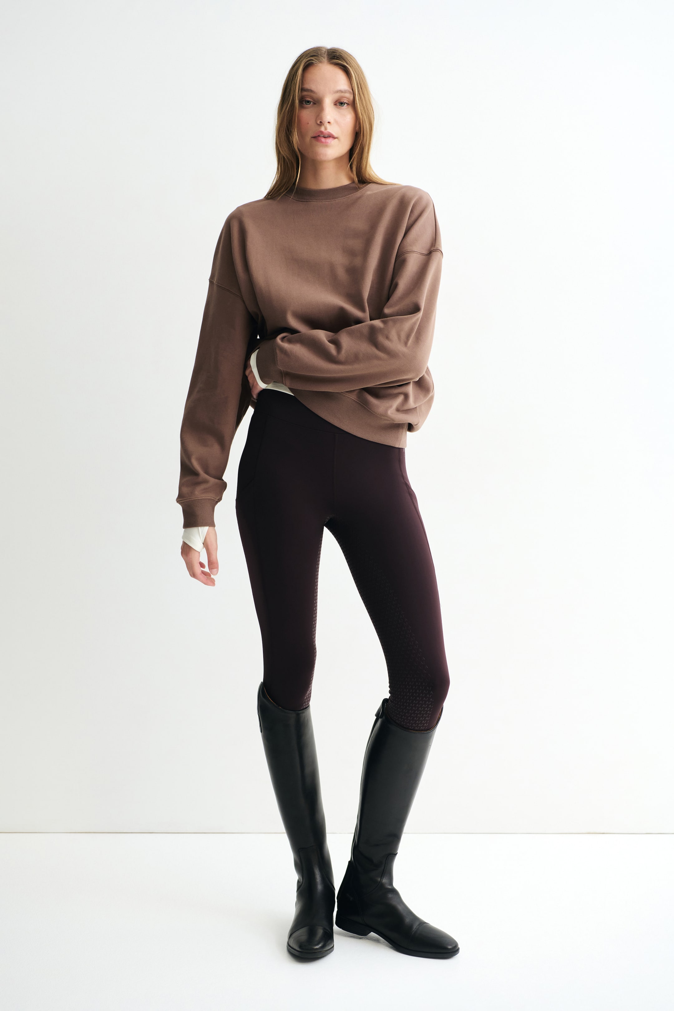 Visualizza l'immagine ingrandita: Una felpa activewear marrone è mostrata frontalmente, con scollo rotondo, spalle scese e dettagli a coste su colletto, polsini e orlo. La felpa è indossata sopra un top bianco a maniche lunghe, con i polsini visibili, ed è abbinata a leggings scuri e stivali neri alti.