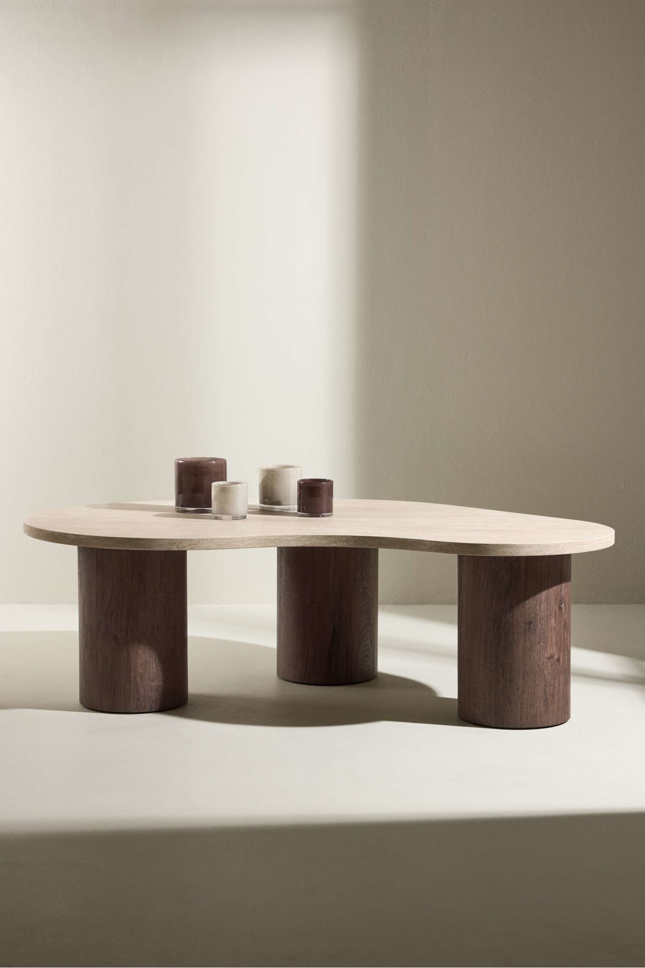 Visualizza immagine più grande: Un tavolino da caffè si presenta con un piano in legno organico e di tonalità chiara, che mostra una venatura visibile, sostenuto da tre spesse gambe cilindriche in legno di tonalità scura. Il tavolo è angolato dalla parte anteriore destra, mettendo in risalto il suo design unico e il contrasto dei materiali.