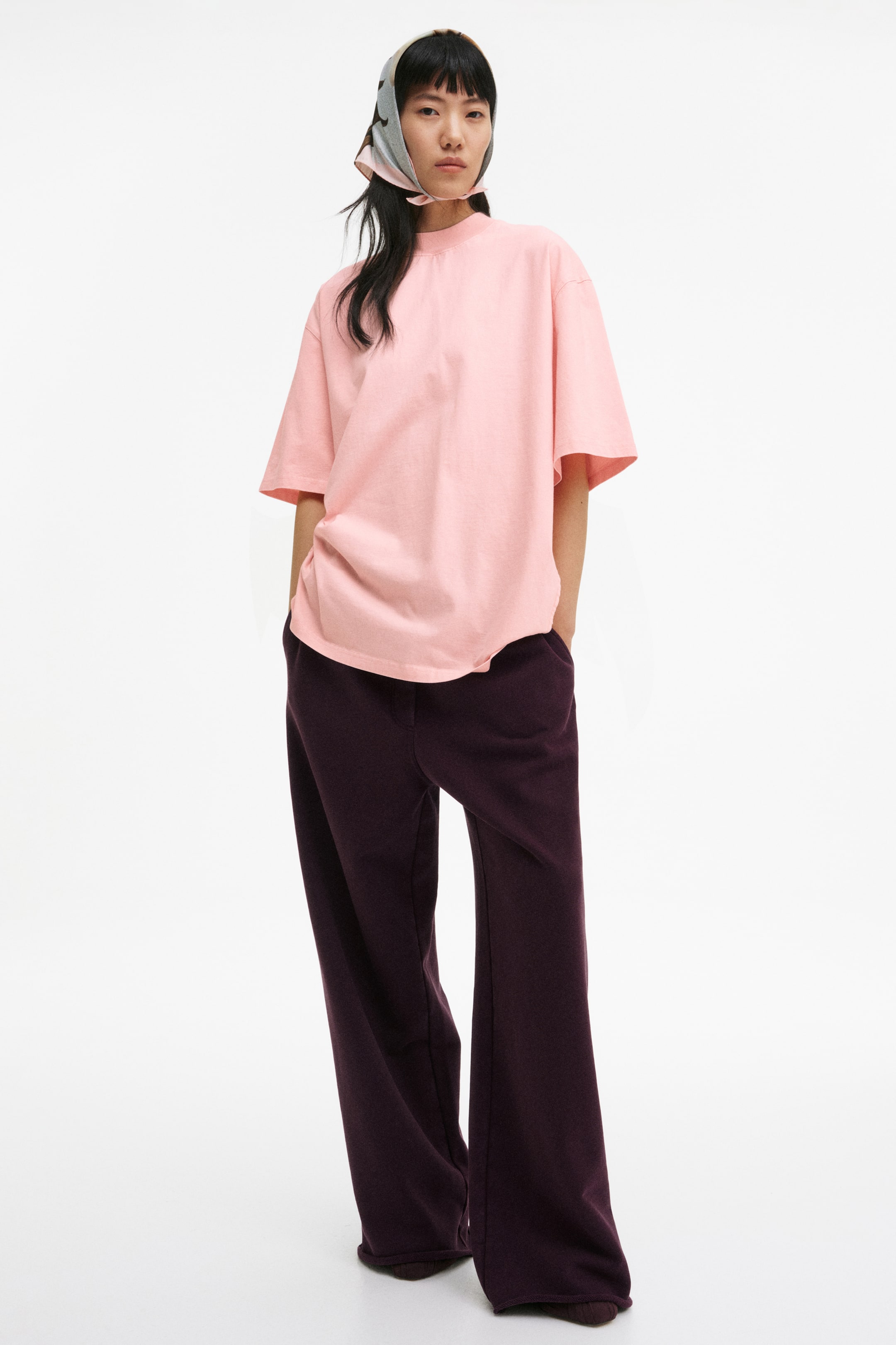 View larger image: The model is wearing an oversized pink t-shirt with a ribbed crew neck, dropped shoulders, and elbow-length sleeves. It features a straight-cut hem and subtly forward-facing, exposed side seams, styled untucked over dark trousers.