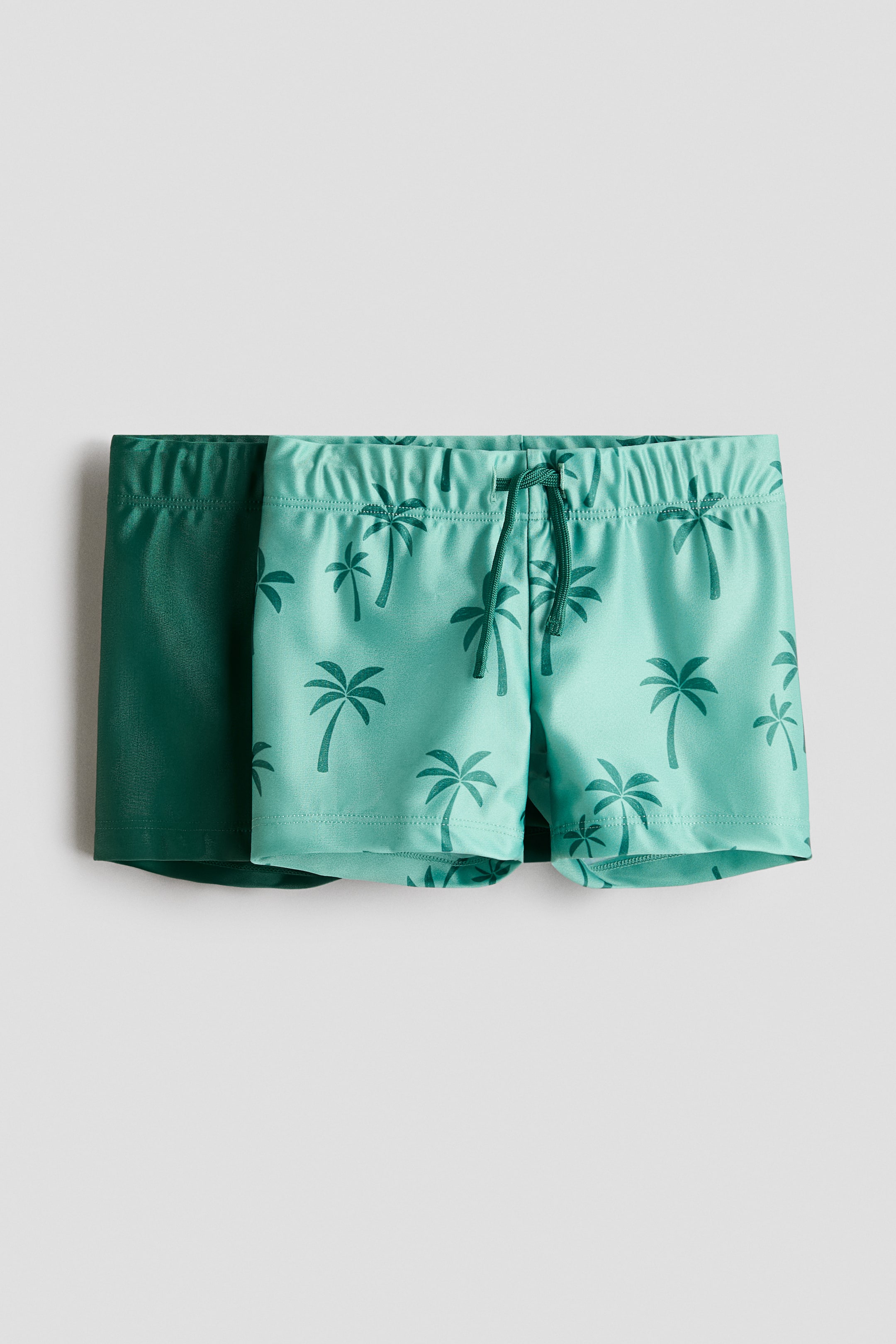 Agrandir l'image: Deux paires de maillots de bain sont présentées de face. La première paire est d'un turquoise clair, ornée d'un imprimé de palmiers vert foncé et dotée d'une taille élastiquée avec un cordon de serrage visible. La deuxième paire, partiellement positionnée derrière, est d'un vert foncé uni avec une taille élastiquée.