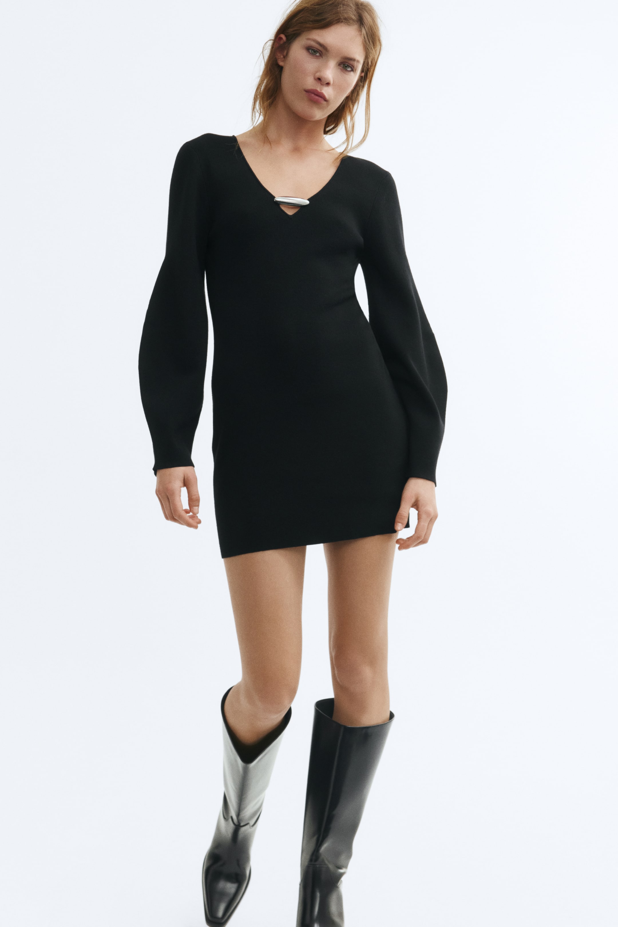 View larger image: The model is wearing a short, fitted black knit dress featuring a deep V-neckline with a horizontal silver metal detail and long, voluminous sleeves with tapered cuffs. The dress is styled with a pair of knee-high black leather boots.