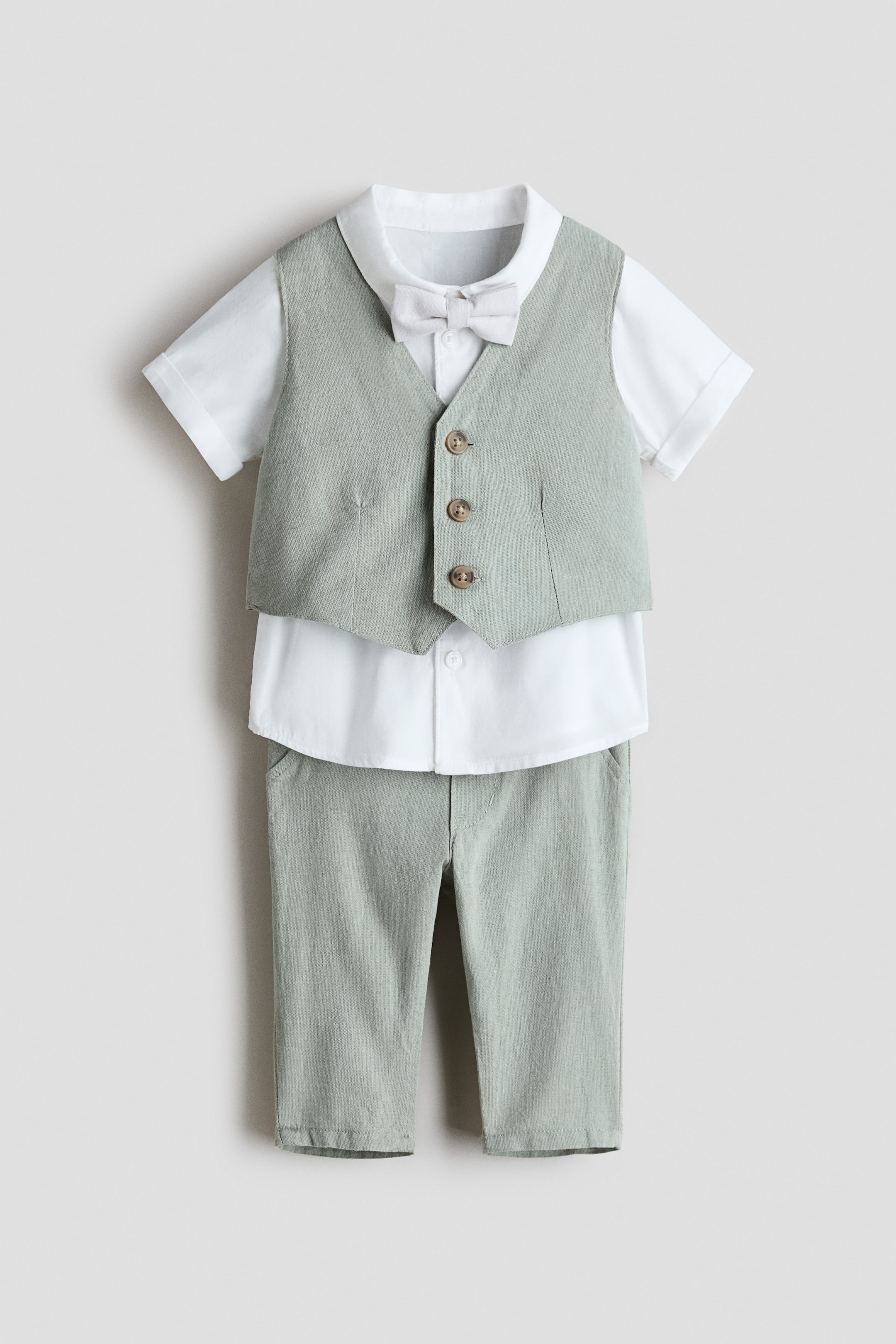Ver imagen más grande: Vista frontal de un conjunto de vestir para bebé que presenta un chaleco de mezcla de lino en verde claro y pantalones a juego, una camisa blanca de popelín de algodón y una pajarita de color claro preanudada. El chaleco muestra tres botones marrones en la parte delantera, los pantalones presentan bolsillos delanteros y la camisa de manga corta incorpora un cuello vuelto y puños con vuelta cosida.