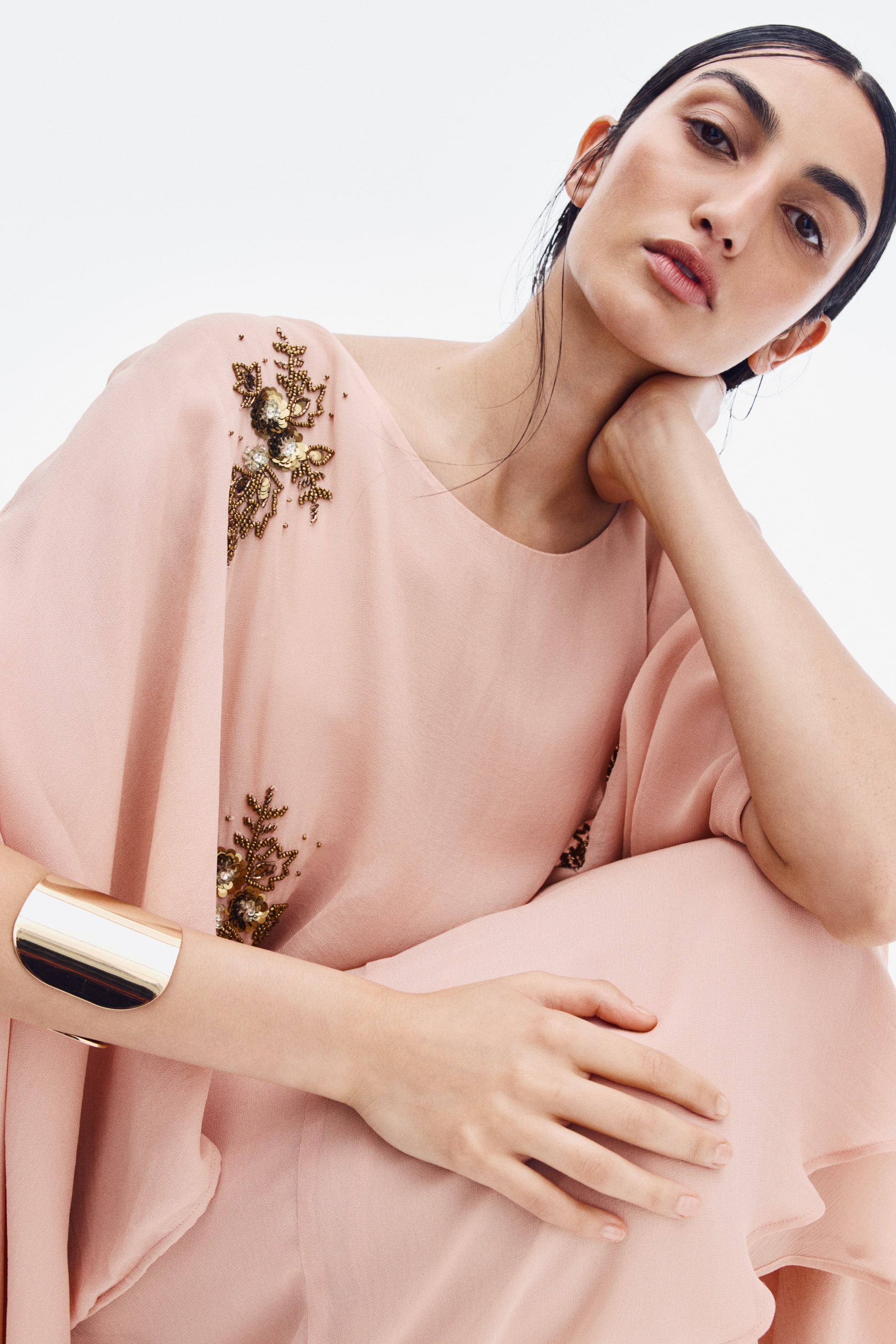 View larger image: A light pink, loose-fit top featuring a round neckline, draped cape sleeves, and gold floral bead embellishments is being worn by the model. The model is posing with one hand resting on their knee, accessorized with a wide gold cuff bracelet on their wrist.