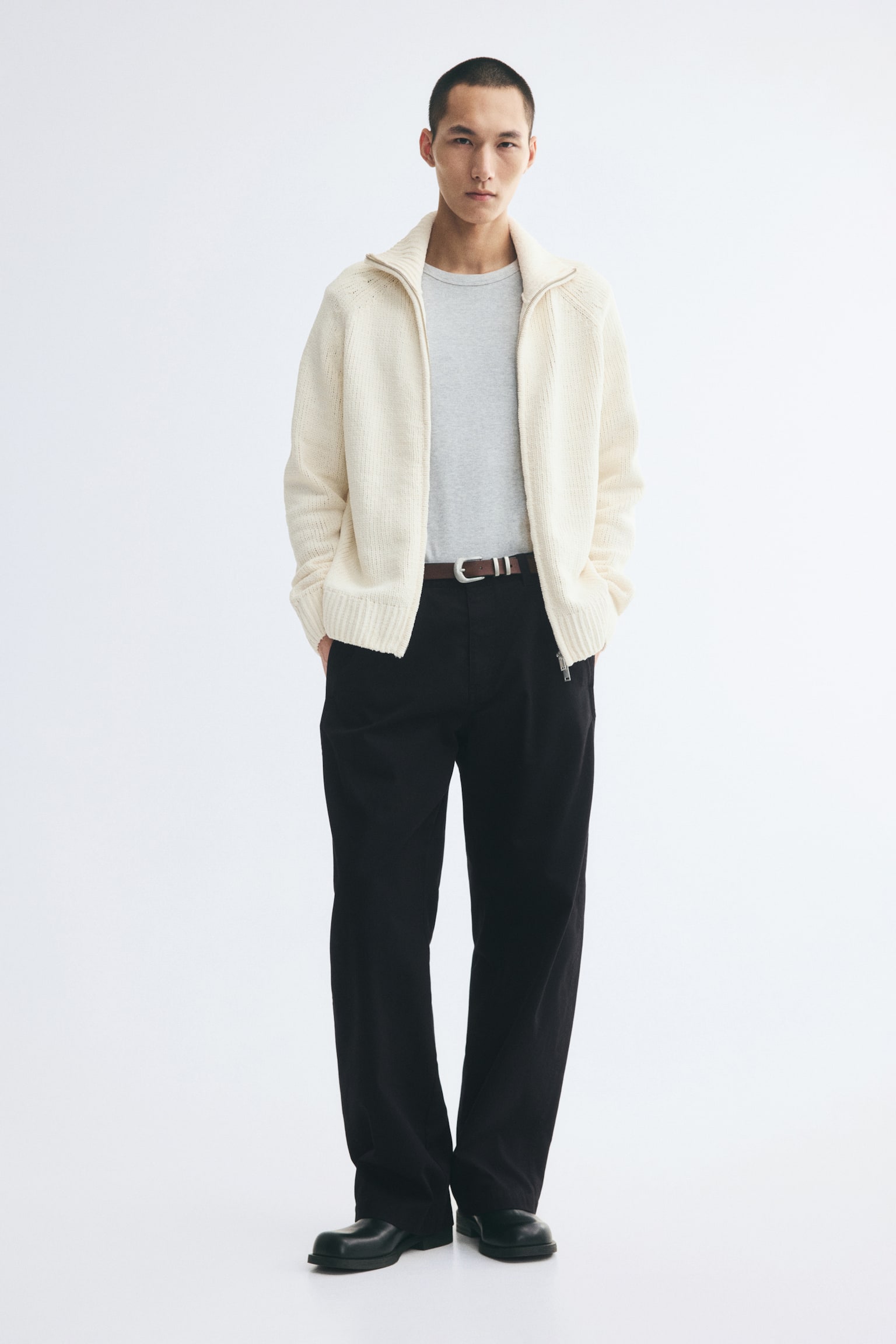 Black cotton twill chinos in a relaxed fit with barrel-shaped legs are worn by the model, who is standing with hands tucked into the side pockets. The trousers are styled with a brown leather belt, a grey t-shirt, an open off-white knit cardigan, and black shoes. - 1