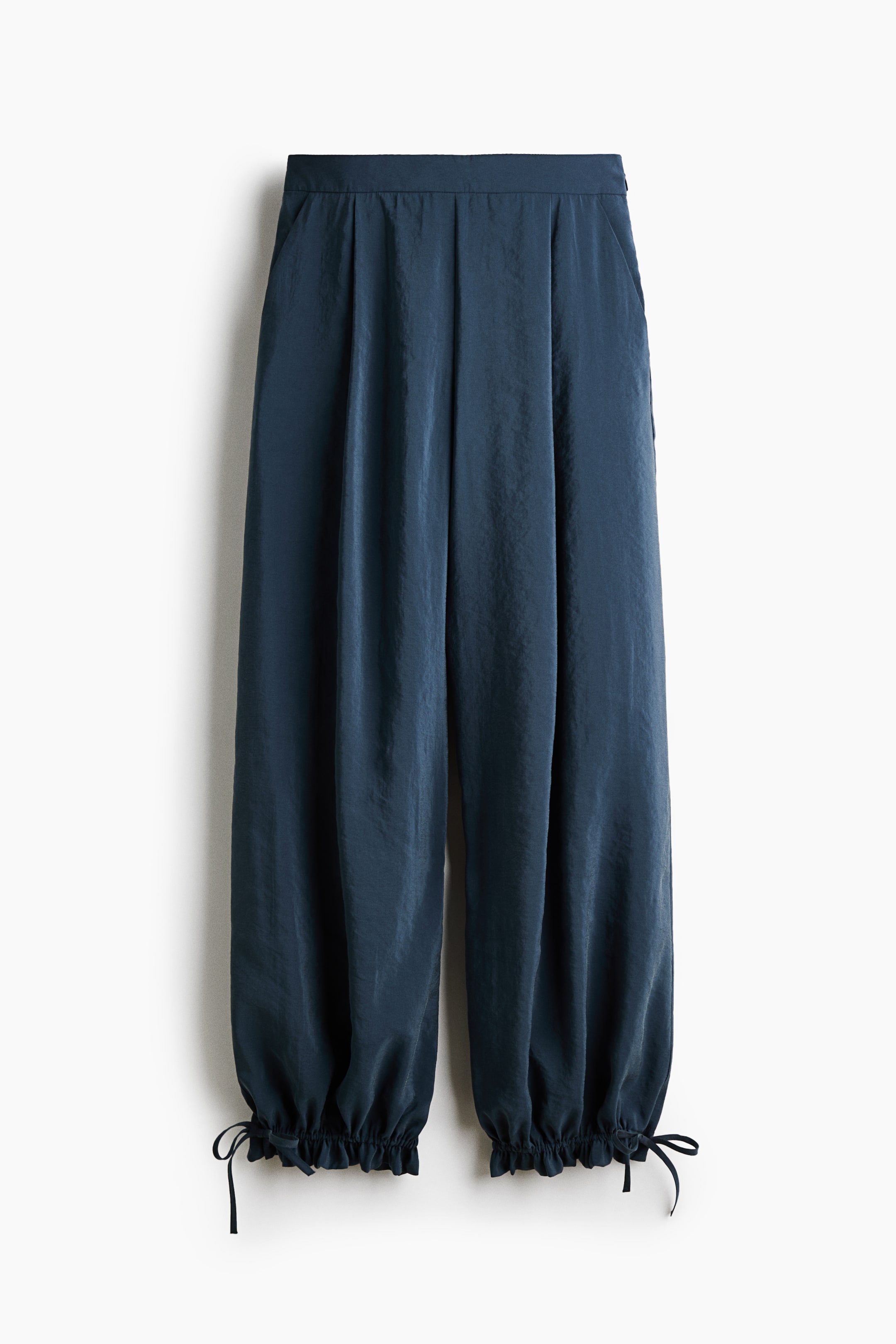 View larger image: These front-facing, dark teal pants hang straight, showcasing a high waist with structured pleats that create a voluminous balloon-leg silhouette. The wide legs taper down to gathered, ruffled cuffs at the ankles, which feature adjustable self-tie drawstrings on the outer sides.