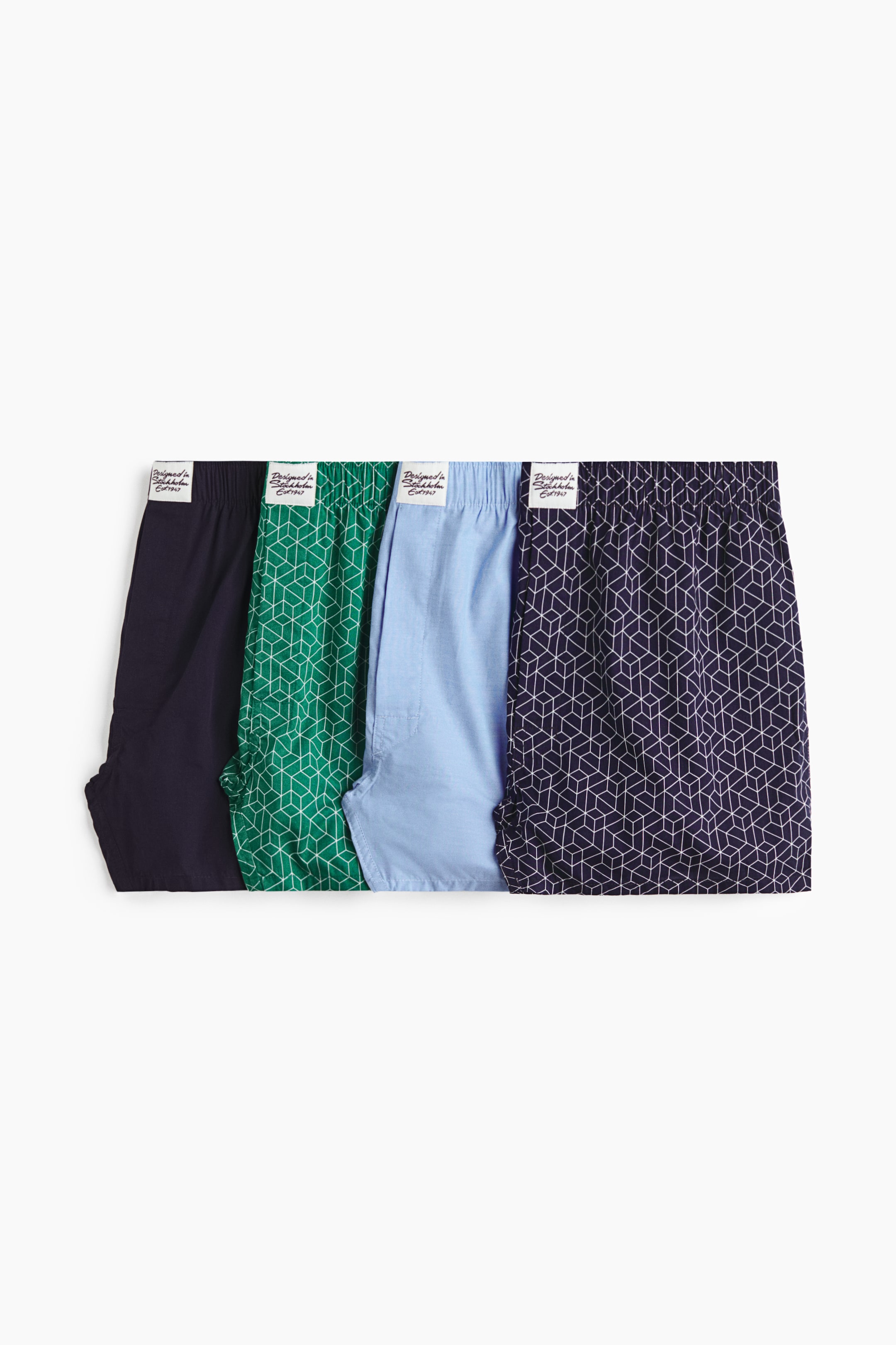 View larger image: Four pairs of men's boxers are displayed side by side, showing their elasticated waistbands with small white brand labels and upper leg sections. The styles include solid navy blue, green with a white geometric cube pattern, solid light blue, and dark blue with a white geometric cube pattern.