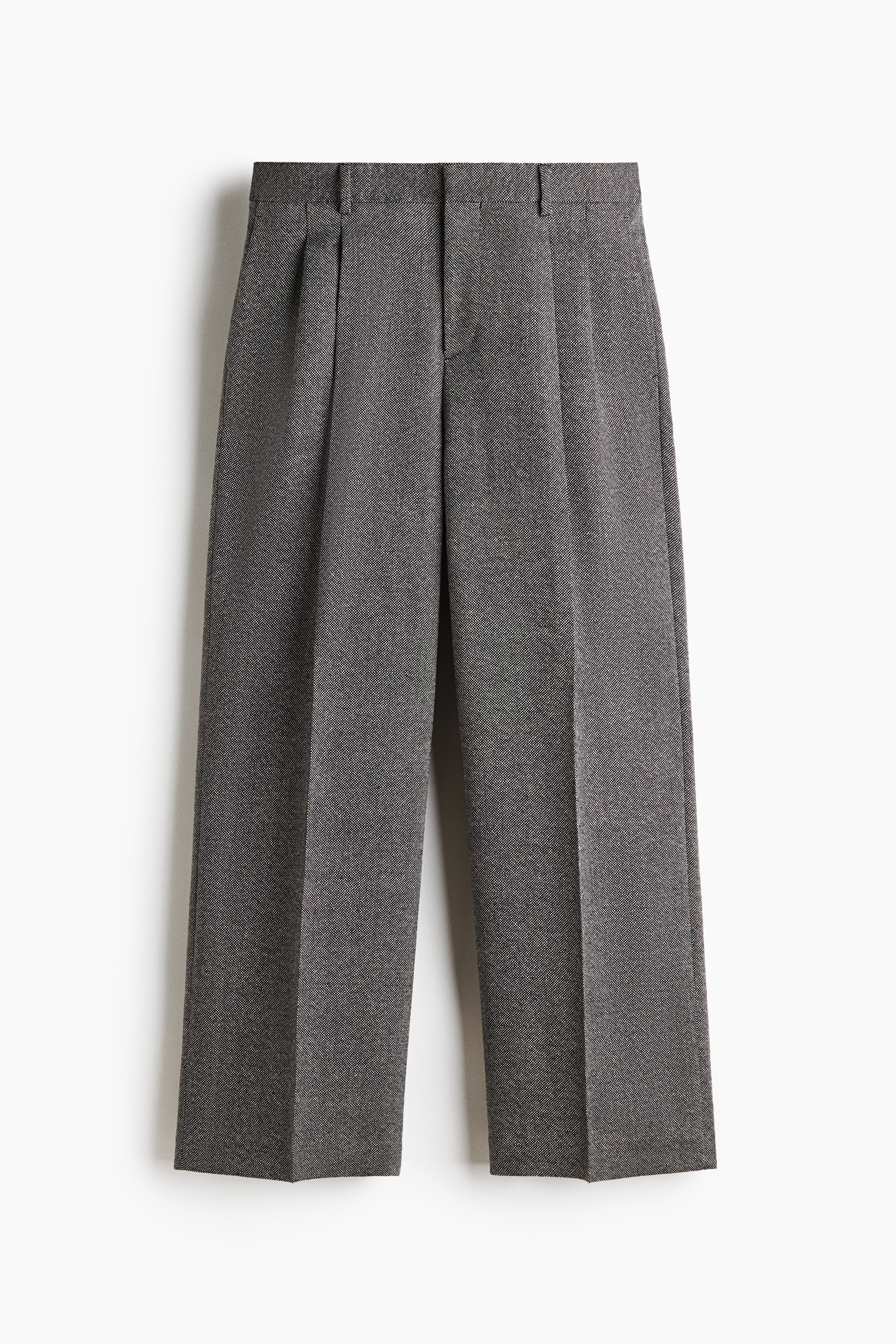 Grey tailored wool-blend trousers featuring a relaxed fit, front pleats, and centre creases are being worn by the model, who is standing with hands in the side pockets. The trousers are styled with a black belt featuring silver-toned metal details, a matching grey zip jacket, a black turtleneck jumper, and black shoes.