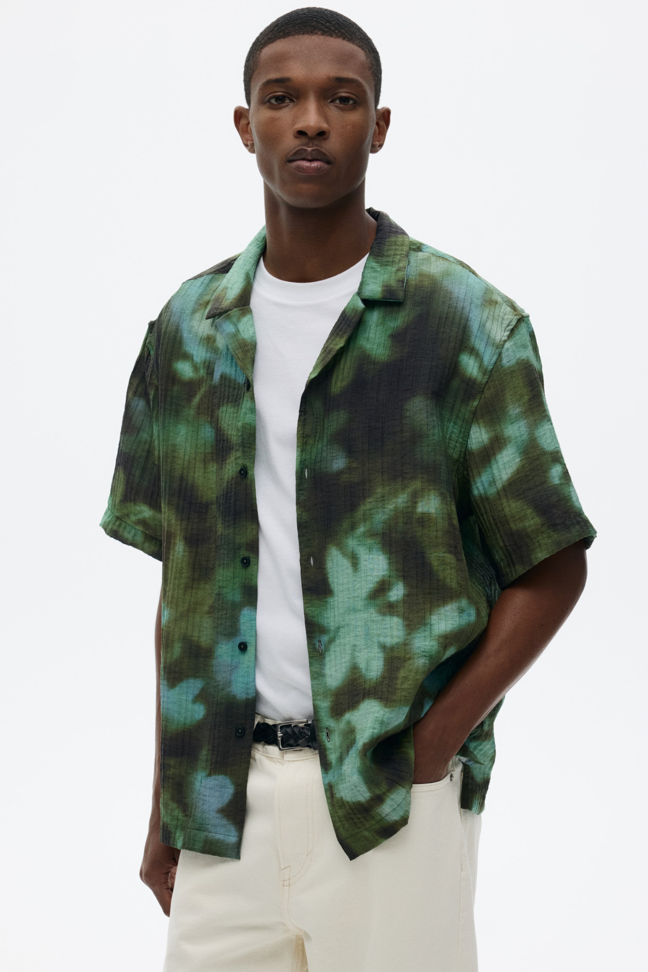 View larger image: The short-sleeved shirt features a mottled green and black abstract pattern with a ribbed texture and a camp collar. It is worn open over a white T-shirt, highlighting its relaxed fit.