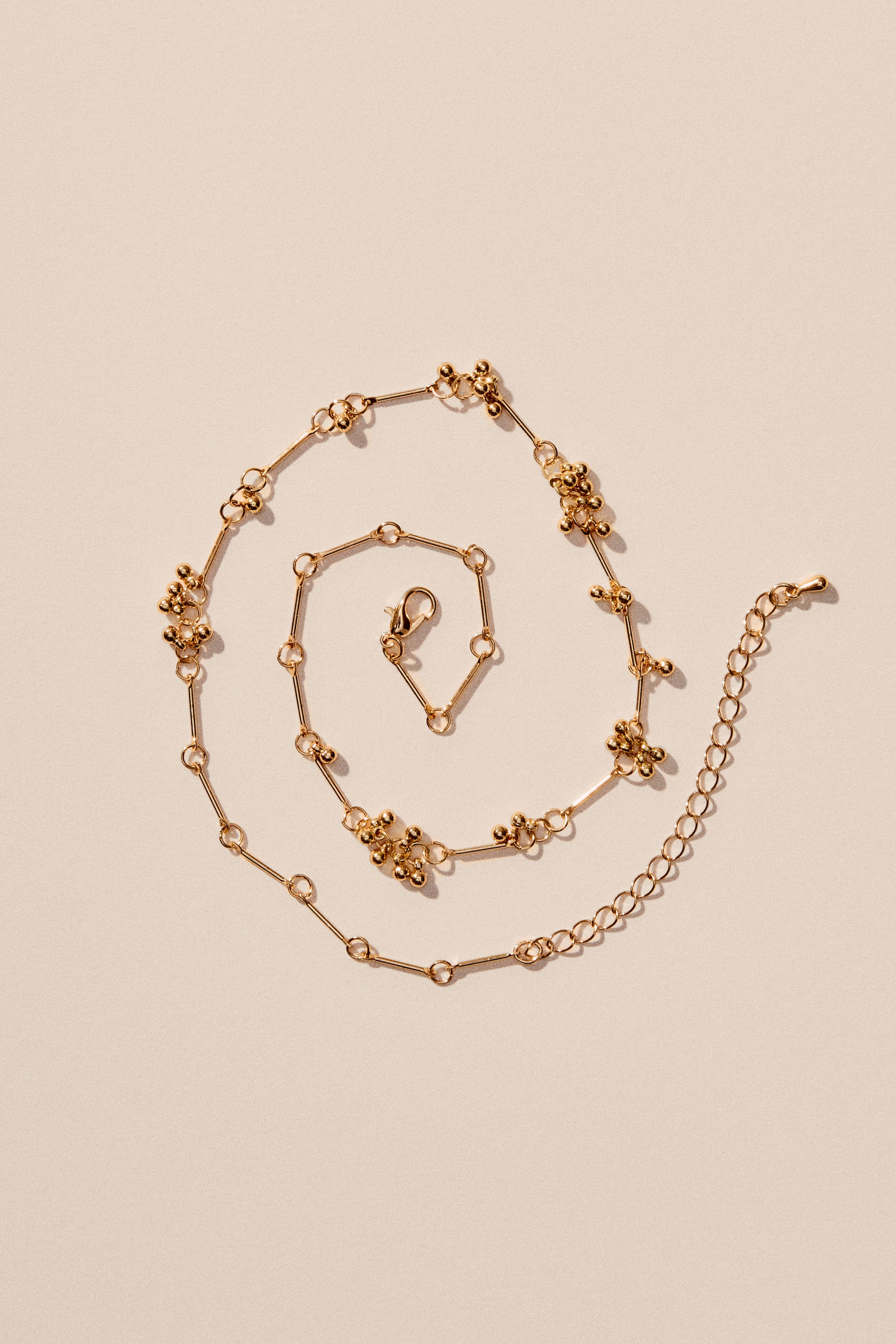 Agrandir l'image: Un collier est conçu à partir d'une chaîne à maillons en métal de couleur dorée, ornée de petits maillons circulaires et de groupes intermittents de petites perles métalliques. Une chaîne d'extension réglable y est attachée, se terminant par un fermoir à mousqueton.