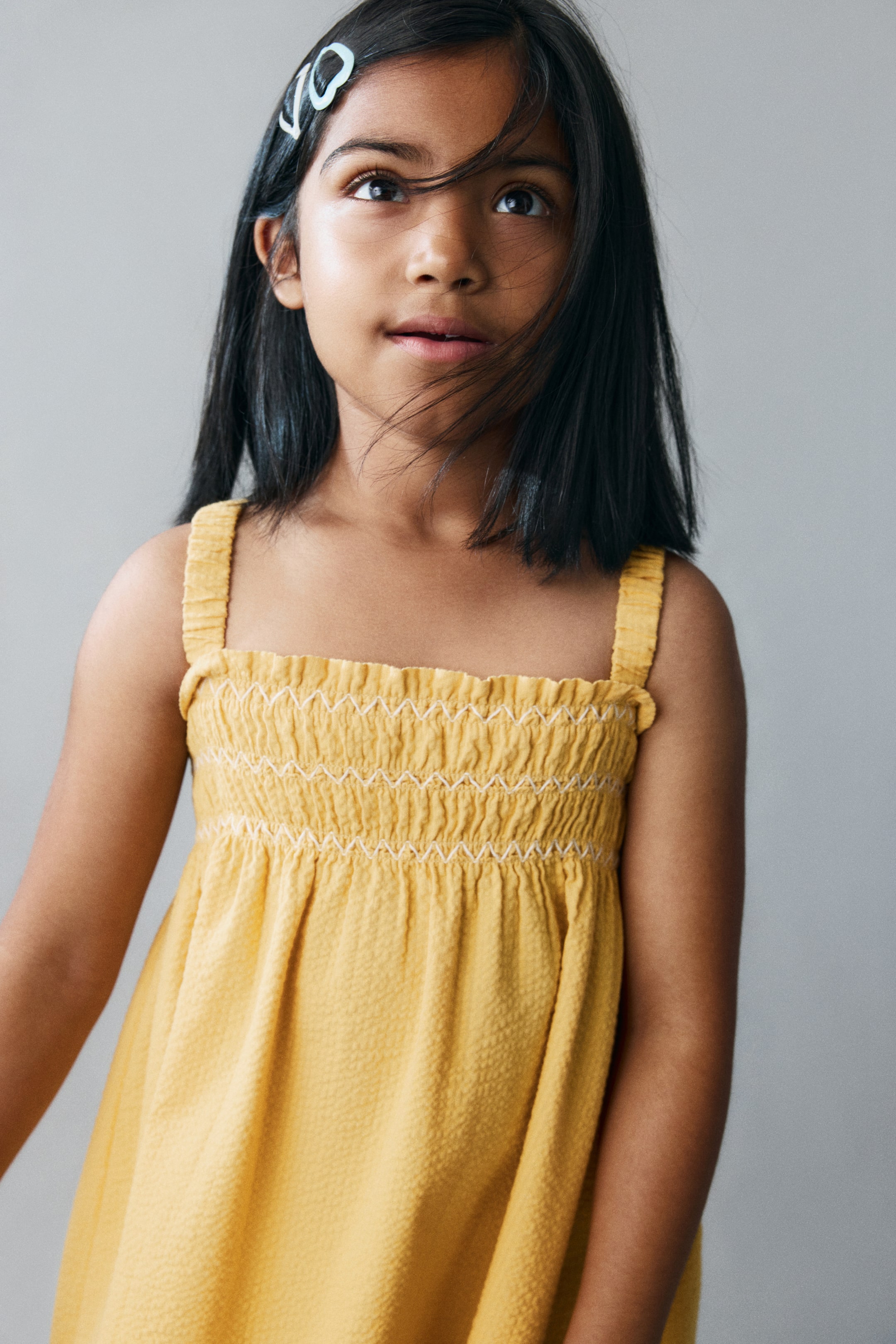 View larger image: A yellow seersucker dress is shown from the front, featuring a smocked bodice with white zigzag embroidery, a frill-trimmed square neckline, and gathered shoulder straps.