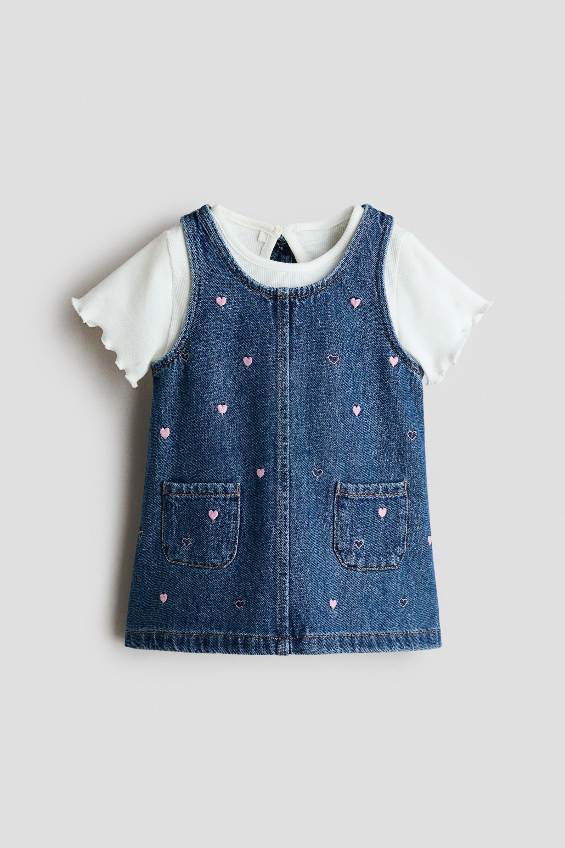 View larger image: A front-facing baby’s dark blue denim pinafore dress is layered over a white short-sleeved ribbed T-shirt. The pinafore features scattered pink embroidered hearts and two front patch pockets, each also displaying a pink embroidered heart, with the white T-shirt showing a ribbed texture and lettuce edge cuffs.