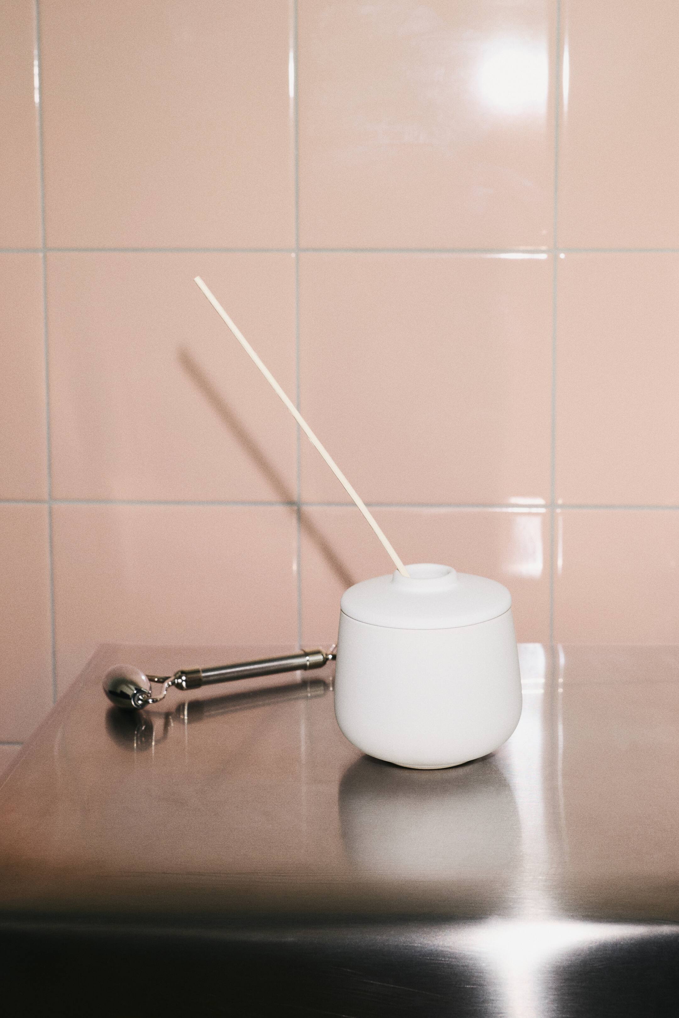 View larger image: A matte white stoneware diffuser featuring a rounded vessel and a matching lid is shown from the front. A single light-coloured wooden reed extends diagonally from the central opening in the lid.