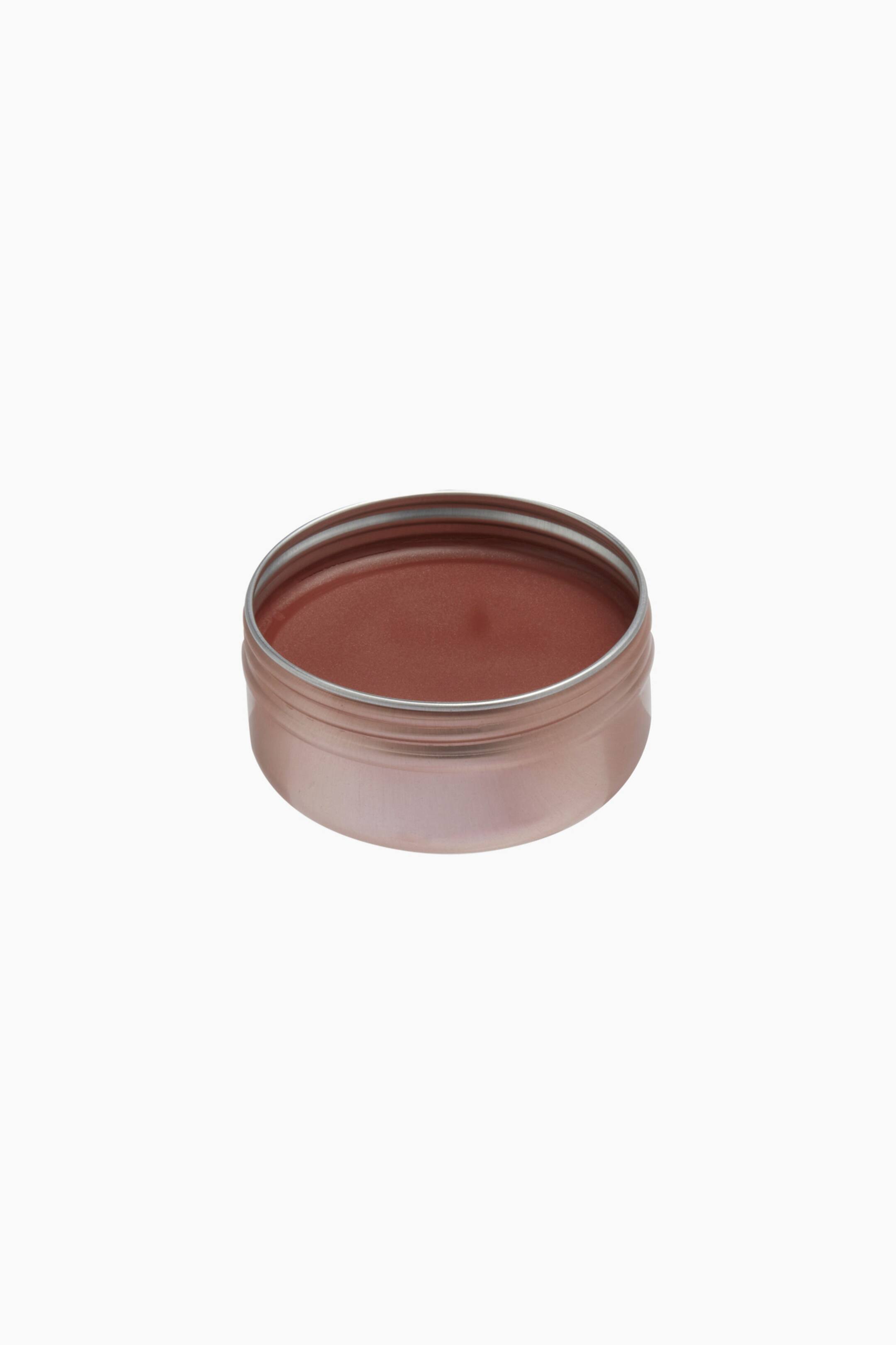 Größeres Bild ansehen: Eine runde, roségoldene Metalldose mit einer glatten, warmen rötlich-braunen Creme-Balm liegt in einer hochwinkligen Dreiviertelansicht. Der offene Behälter zeigt das feste, leicht glänzende Make-up-Produkt, das bis zum Rand gefüllt ist.