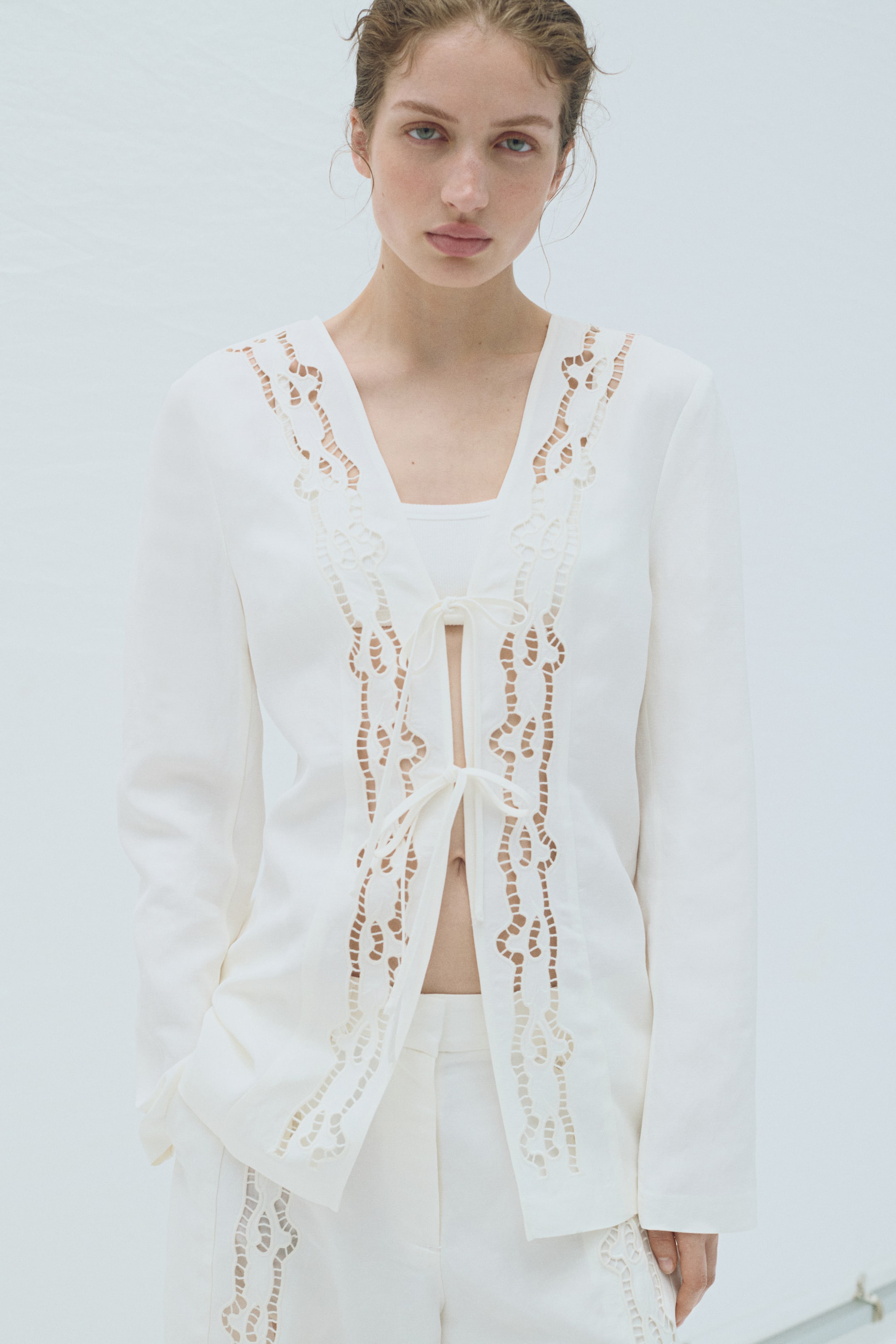 View larger image: The model is wearing a white, collarless blazer featuring ladder lace insets running down its open front. It is fastened at the front with two sets of narrow ties and is styled over a white top.