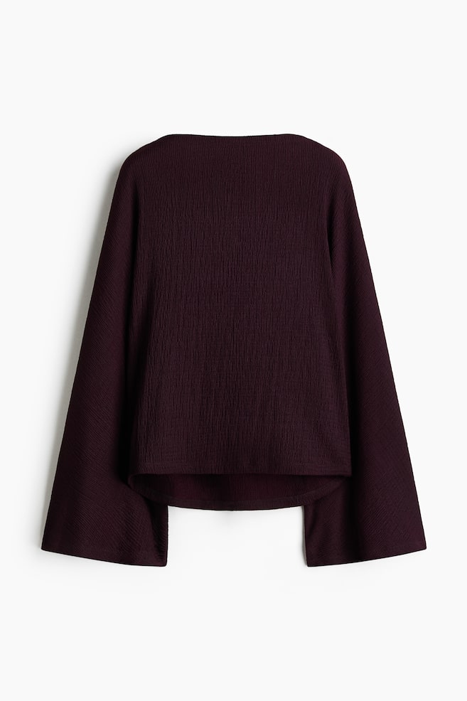 A dark purple textured top is displaying its back view. It is featuring a wide square neckline, wide bell sleeves, and an uneven hem with a central split. - 1