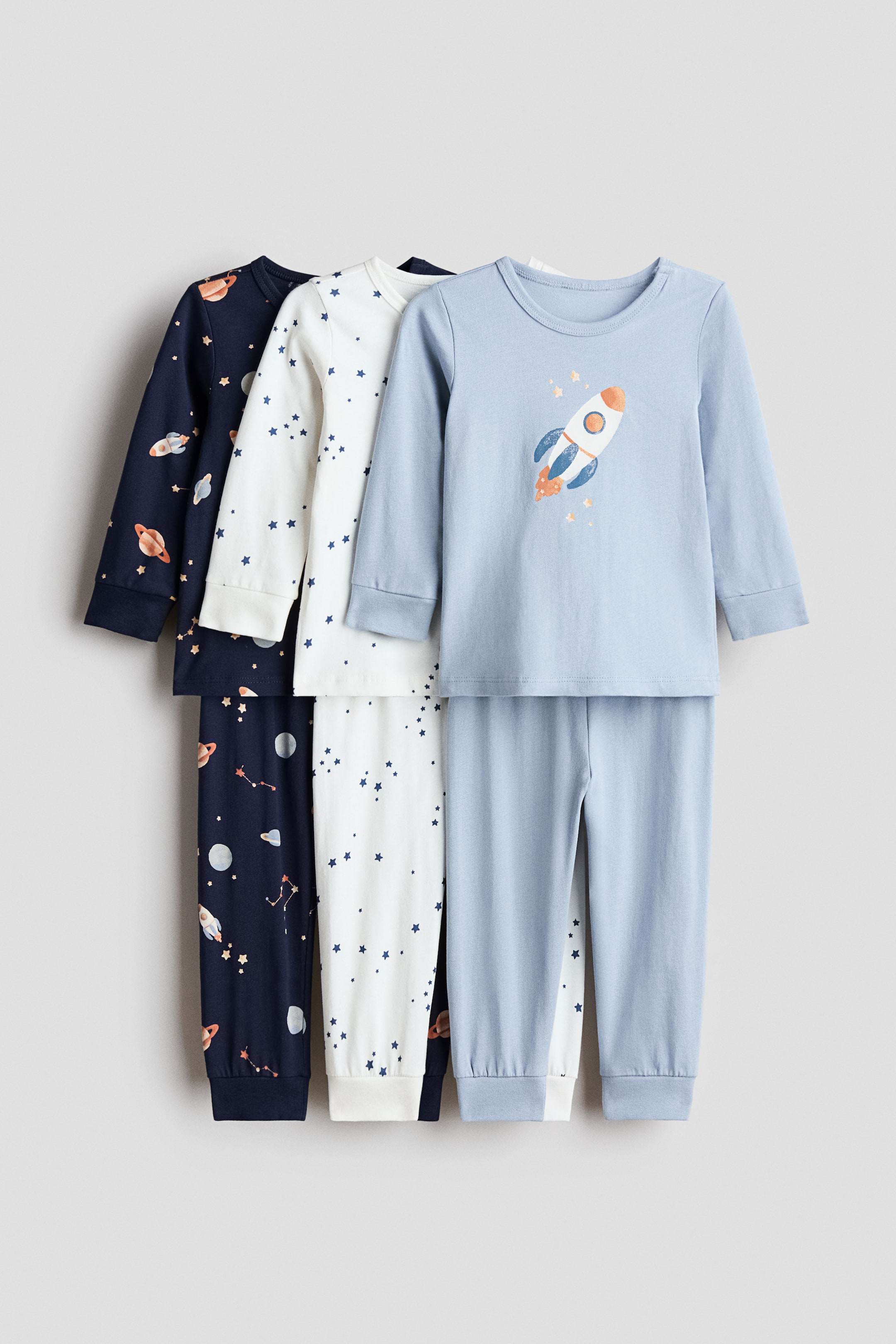 Agrandir l'image: Trois ensembles de pyjamas pour enfants sont exposés, chacun comprenant un haut à manches longues et un pantalon assorti avec des poignets côtelés. Le premier ensemble bleu clair présente un motif de fusée sur le devant du haut, tandis qu'un ensemble blanc cassé derrière est orné d'étoiles bleues, et un ensemble bleu foncé représente divers éléments de l'espace.