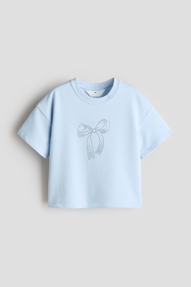 A light blue cropped short-sleeved top is presented from the front, featuring a ribbed crew neck and dropped shoulders. A prominent bow design, made from numerous small rhinestones, is centrally positioned on the front. - 1