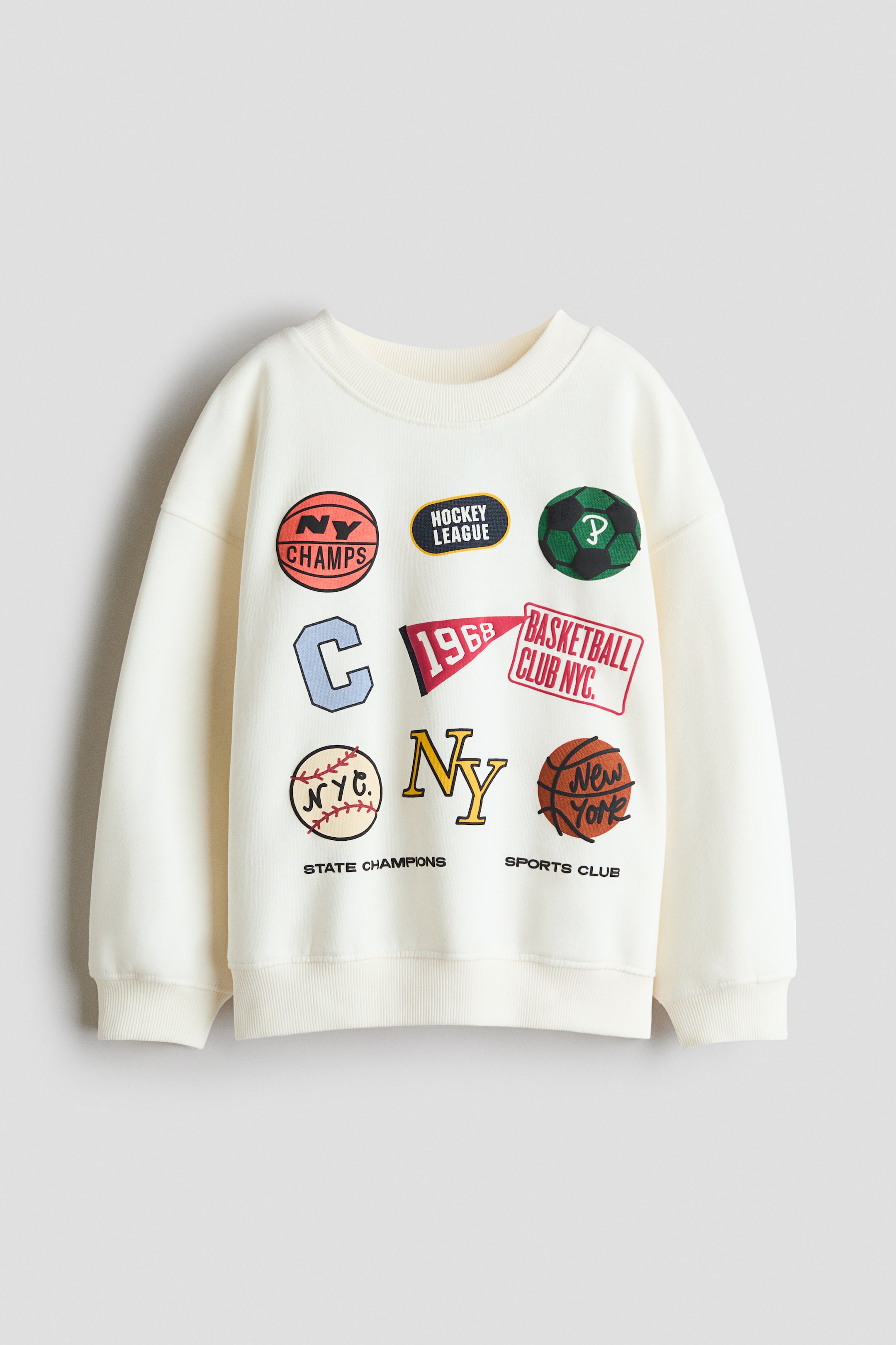 View larger image: A loose-fit top in printed sweatshirt fabric is shown from the front, featuring a round, rib-trimmed neckline, dropped shoulders, and ribbed trims at the cuffs and hem. The cream-coloured top is displaying various colourful sports-themed patches and text, including basketballs, a football, and phrases like 'NY CHAMPS' and 'HOCKEY LEAGUE' on its front.
