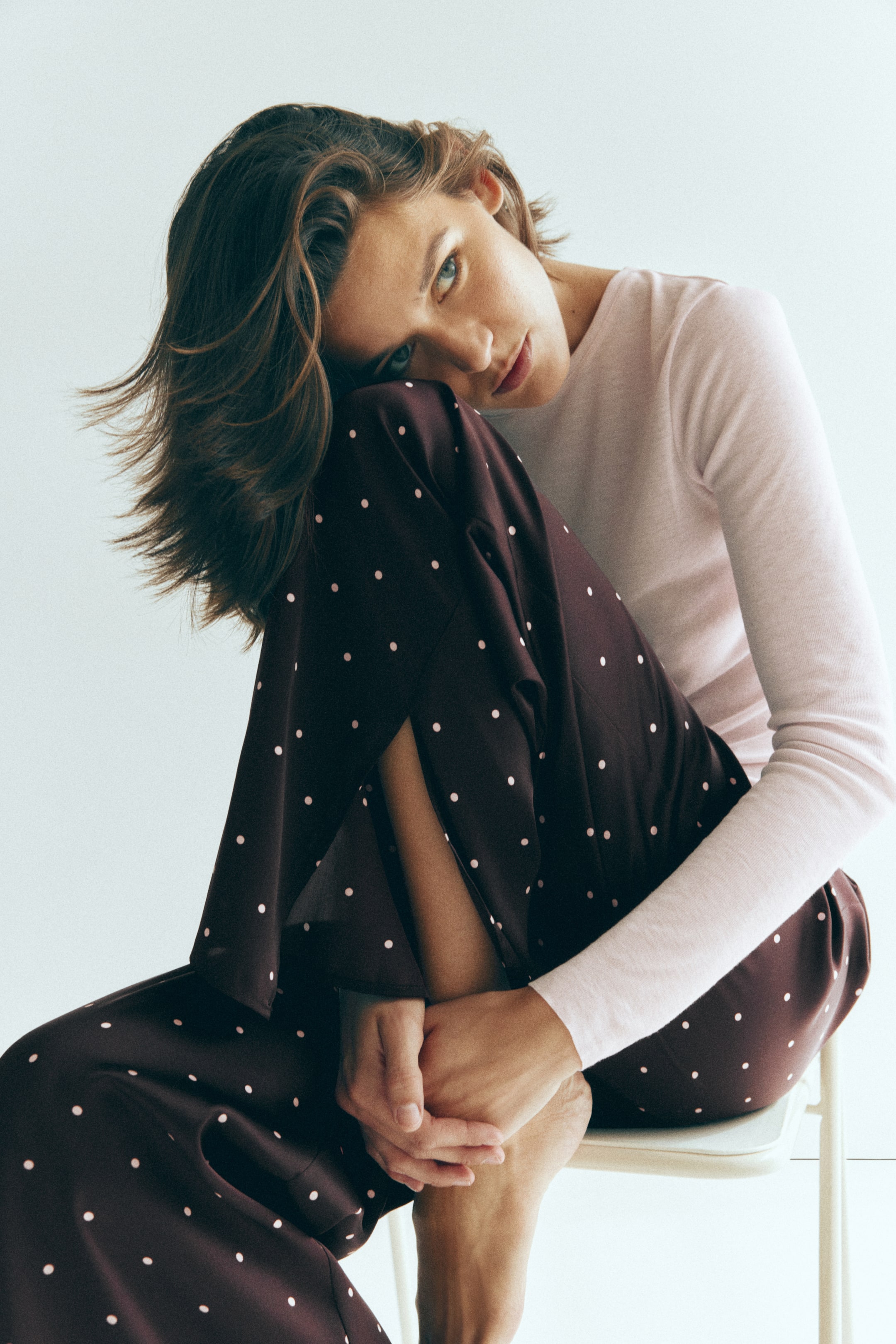 Visualizza immagine più grande: Un completo da pigiama composto da un top a maniche lunghe in costina rosa chiaro e pantaloni in satin marrone scuro a pois viene indossato dalla modella. La modella è seduta con un ginocchio sollevato verso il petto, mettendo in risalto il dettaglio dello spacco nell’orlo dei pantaloni drappeggiati.
