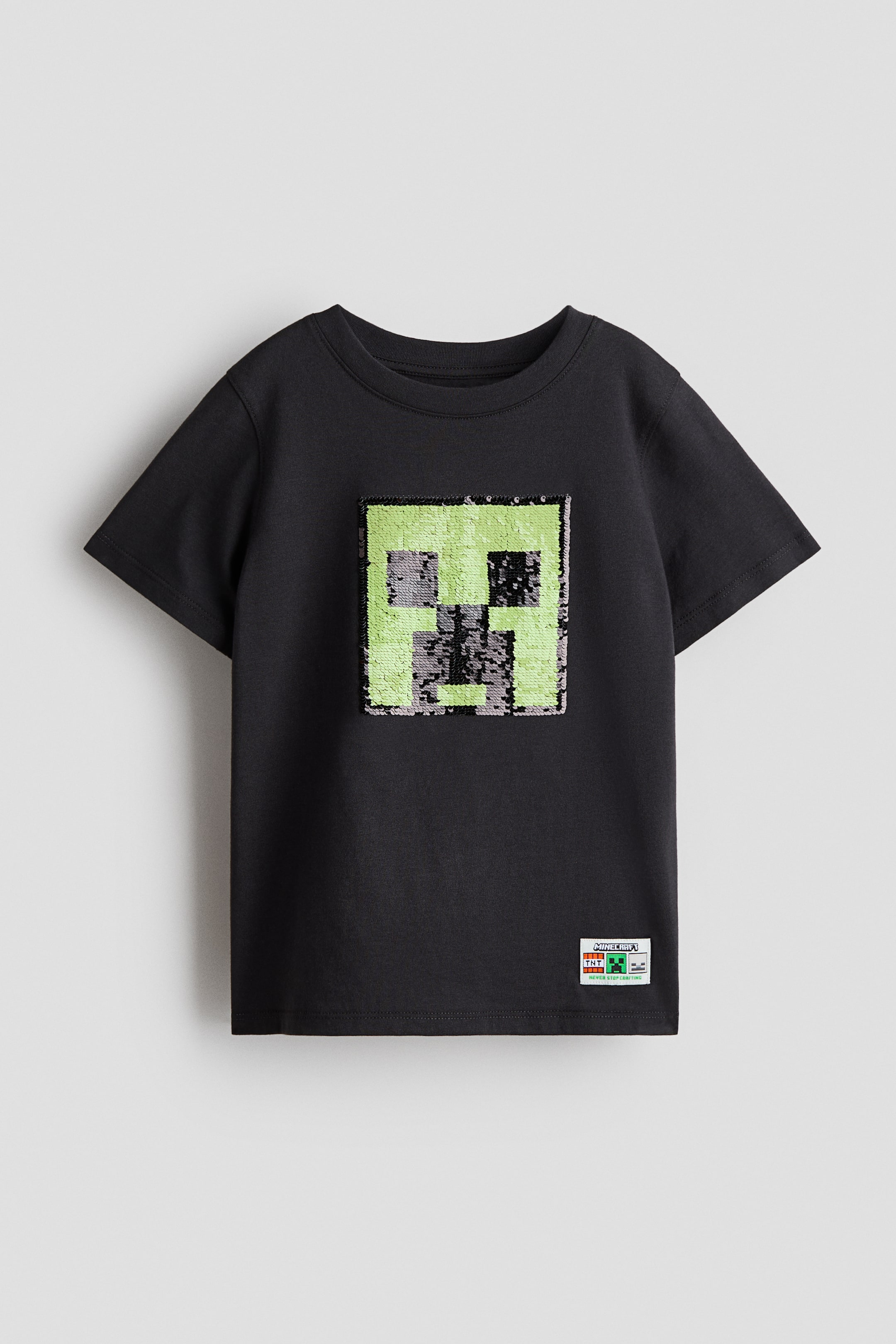 Ampliar la imagen: Una camiseta negra se muestra de frente y presenta un gran diseño cuadrado pixelado de una cara de Creeper en verde y gris, formada por lentejuelas en el pecho. Cuenta con mangas cortas, un ribete acanalado alrededor del cuello y un dobladillo de corte recto.