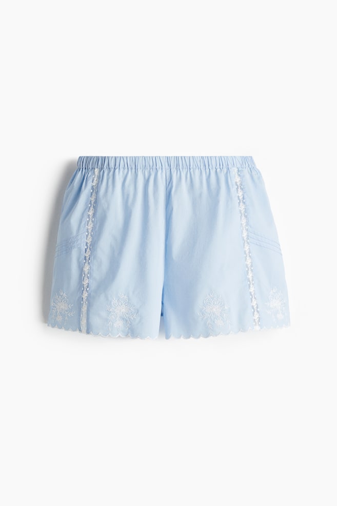 Ein Paar hellblaue Shorts wird aus der Vorderansicht präsentiert, mit einem elastischen Bund, weißer Spitzenverzierung entlang der Seitennähte und weißer Blumenstickerei über den gewellten Säumen. - 1