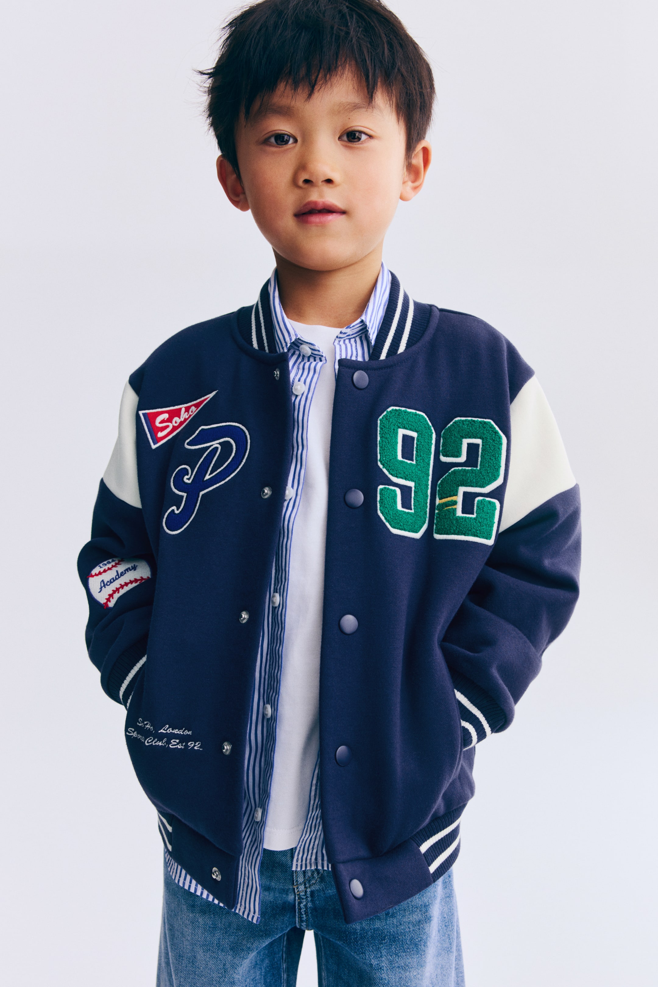 View larger image: A navy blue and white varsity jacket is shown from a front-facing view, featuring a navy body with white sleeves and a striped ribbed collar, cuffs and hem. It showcases snap-button closures and multiple embroidered patches, including a blue "P" and a red "Soho" on the left chest, a green "92" on the right chest, and a baseball graphic on the left sleeve.