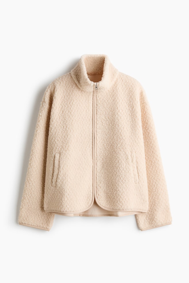 A cream-colored sherpa fleece jacket is shown from the front, featuring a high stand-up collar and a full-length zipper closure. The jacket is designed with long sleeves, vertical welt pockets, and a curved hemline finished with matching tonal binding. - 1
