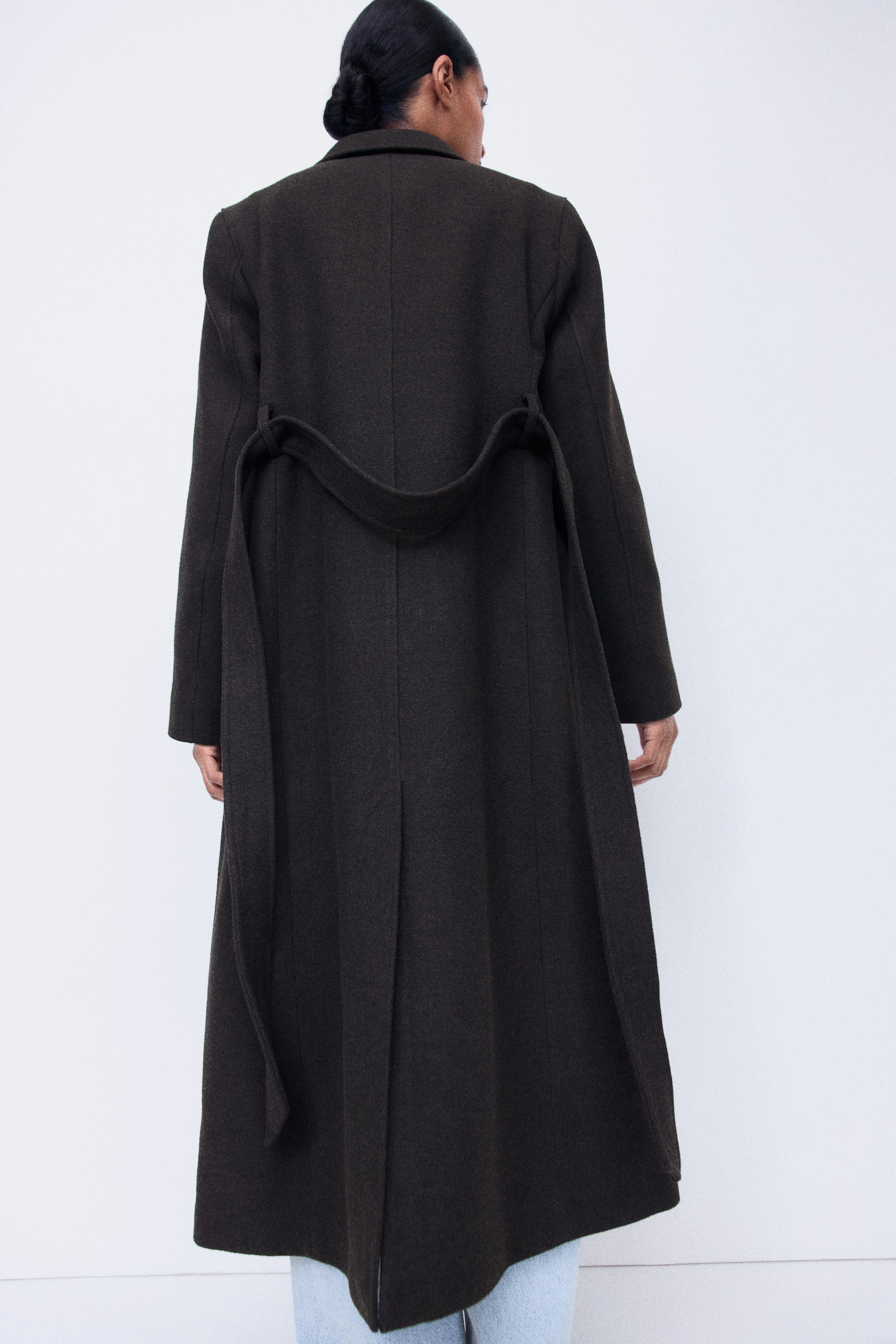 View larger image: A dark olive-green, calf-length woven coat is being worn by the model, shown from the rear. The coat features a matching tie belt draped loosely across the back waist and a single back vent at the bottom hem.