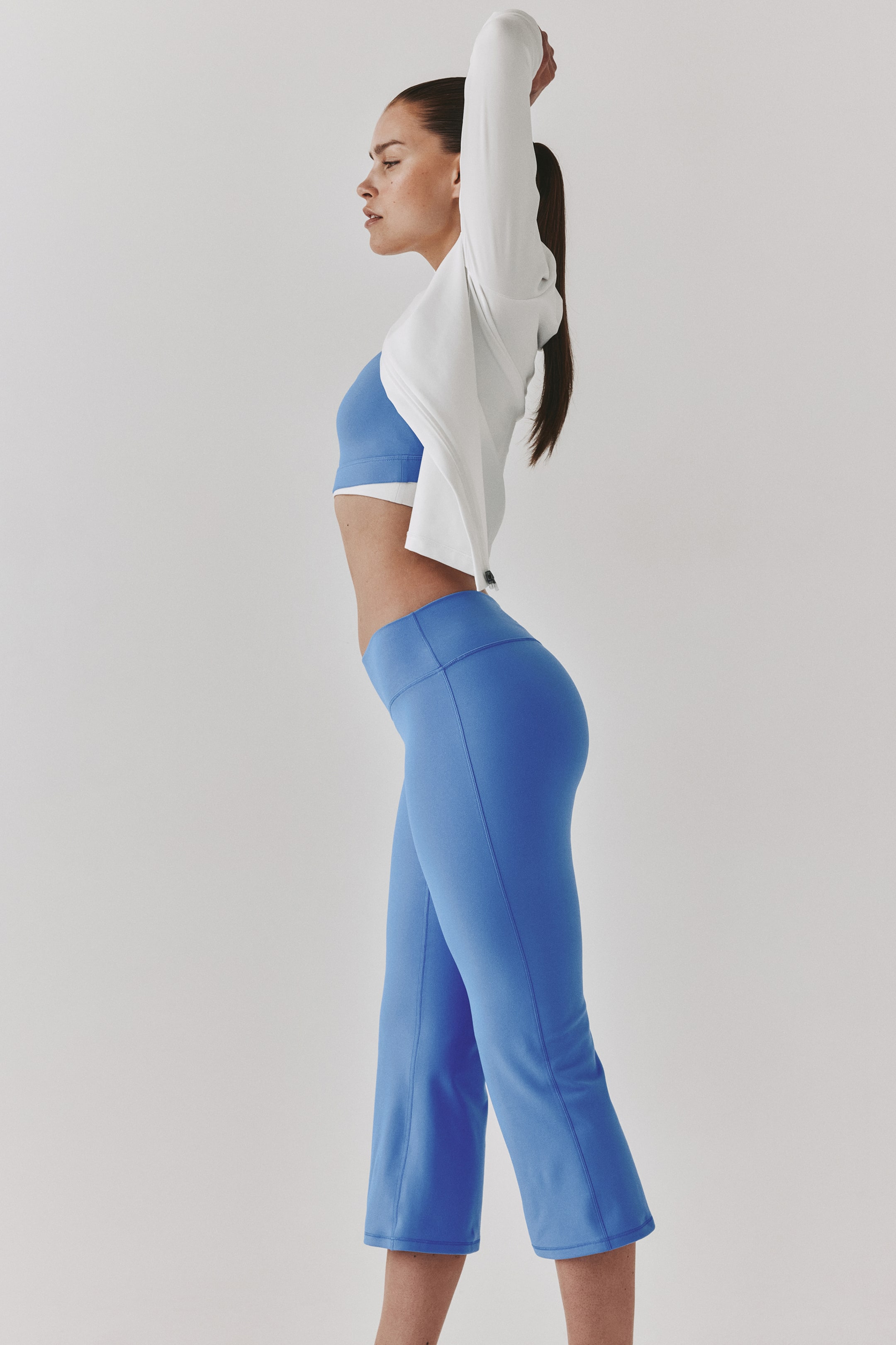 View larger image: The model is wearing a blue sports bra, blue high-waisted cropped flared athletic pants, and a white long-sleeved top. As she stretches with her arms raised, the white top subtly reveals the layered sports bra beneath.