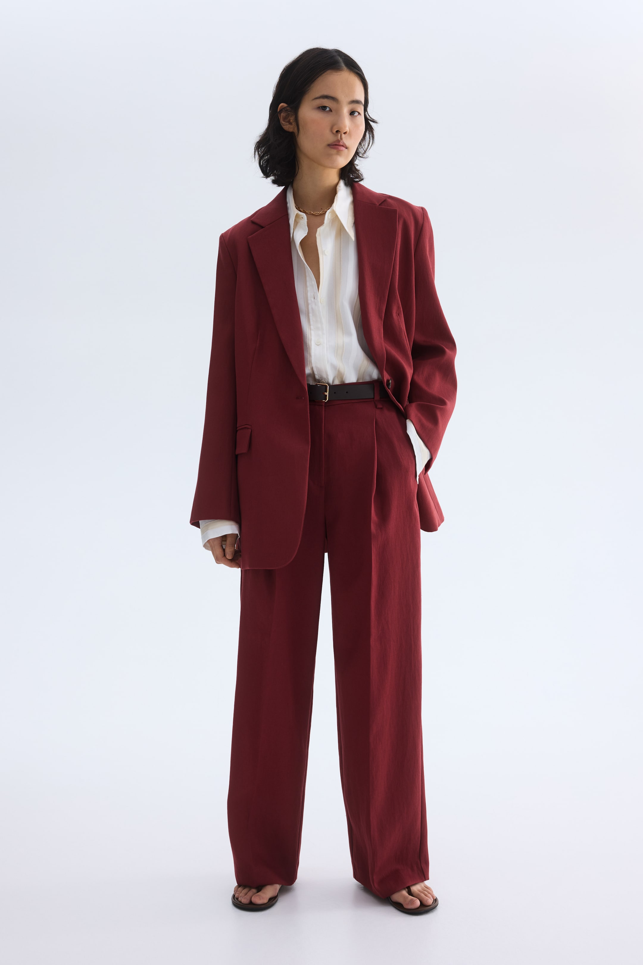 Zobacz większy obraz: Prezentowany jest głęboki czerwony garnitur dwuczęściowy, składający się z oversize'owej marynarki jednorzędowej z klapami w szpic oraz szerokich spodni z zakładkami. Stylizowany jest na jasną koszulę w paski z guzikami i przewiązany w talii ciemnym paskiem.
