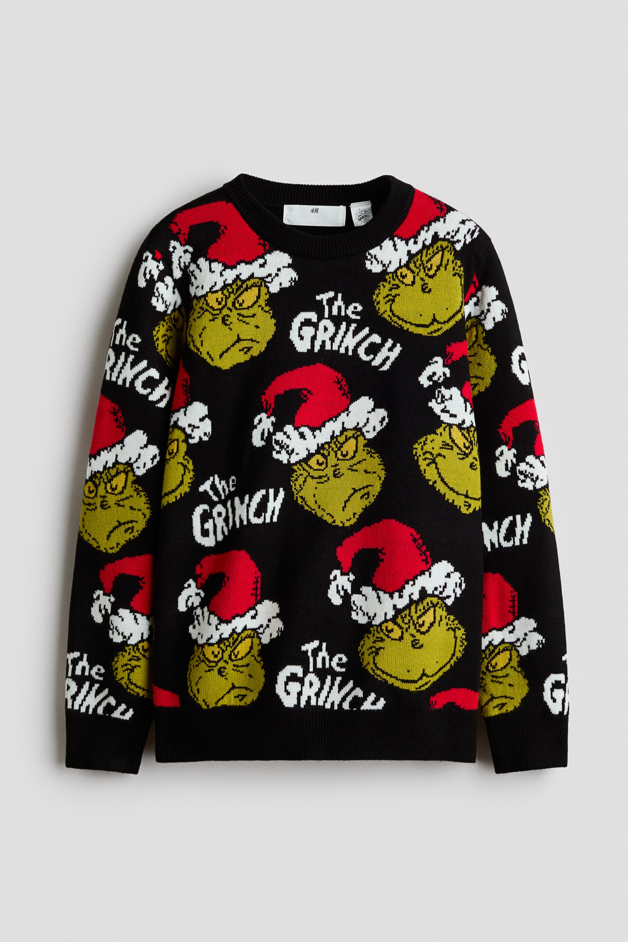 Vis større bilde: En svart strikkegenser sett forfra viser et heldekkende mønster av Grinch med en rød og hvit nisselue, avbrutt av den hvite teksten «The GRINCH». Genseren har en ribbestrikket rund hals, ribbestrikkede mansjetter og en ribbestrikket nederkant.