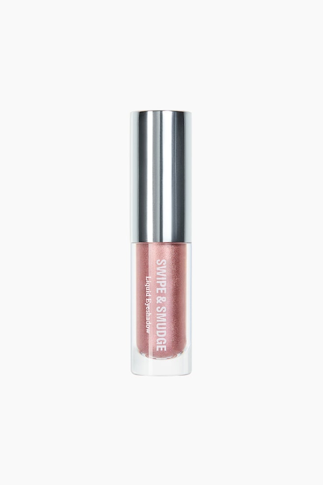 Un fard à paupières liquide est posé verticalement, de face, présentant un flacon cylindrique transparent rempli d'un liquide chatoyant couleur or rose. Il est surmonté d'un bouchon cylindrique argenté et réfléchissant, et les mots 'SWIPE & SMUDGE Liquid Eyeshadow' sont visibles verticalement à l'avant. - 1