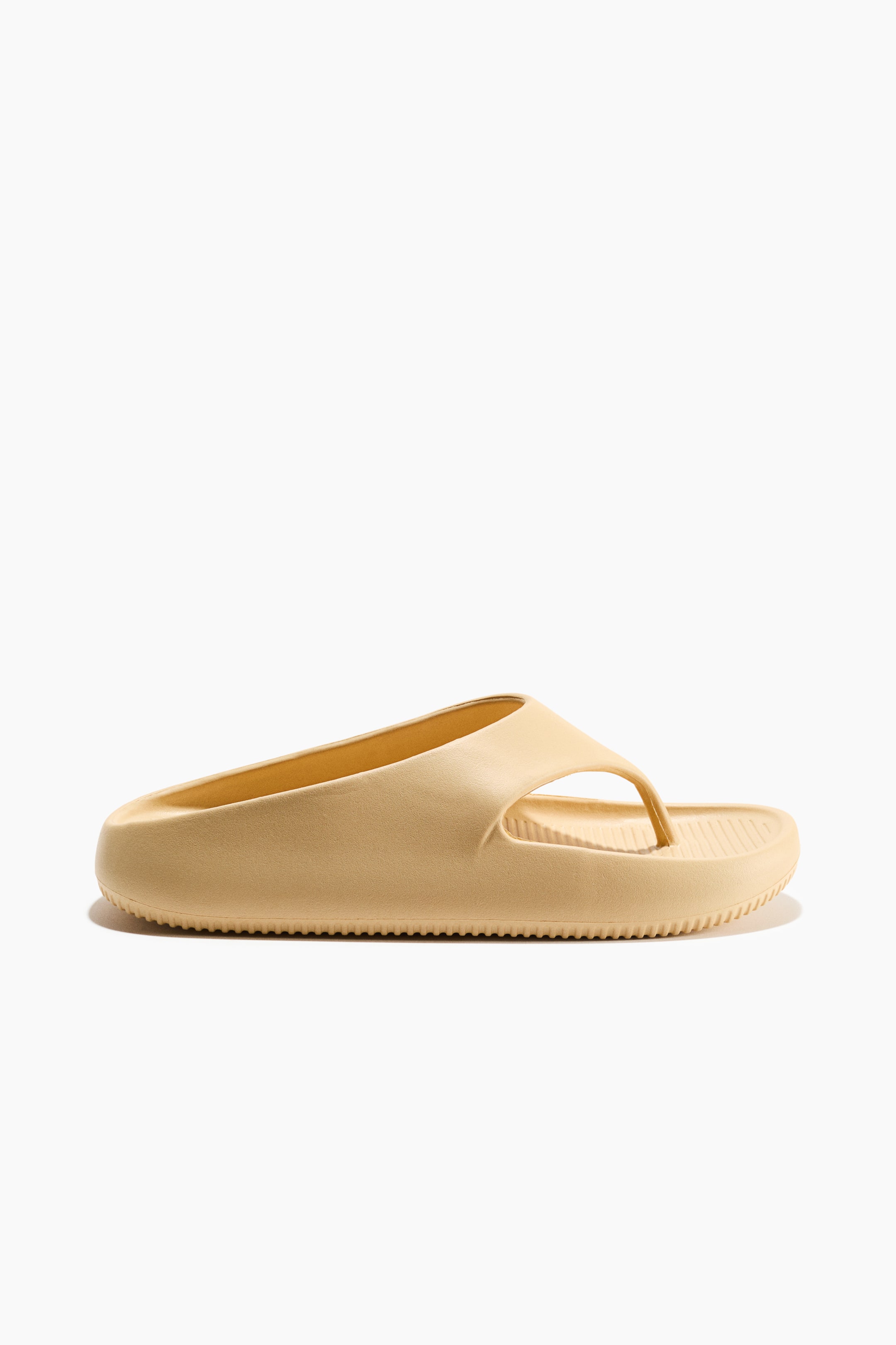Agrandir l'image: Une seule sandale tong beige clair est présentée sous un angle de vue latéral-frontal. Elle se caractérise par une semelle épaisse et moulée avec de subtiles rainures verticales le long du bord et un lit de pied nervuré. Une large bride moulée forme un entre-doigt.