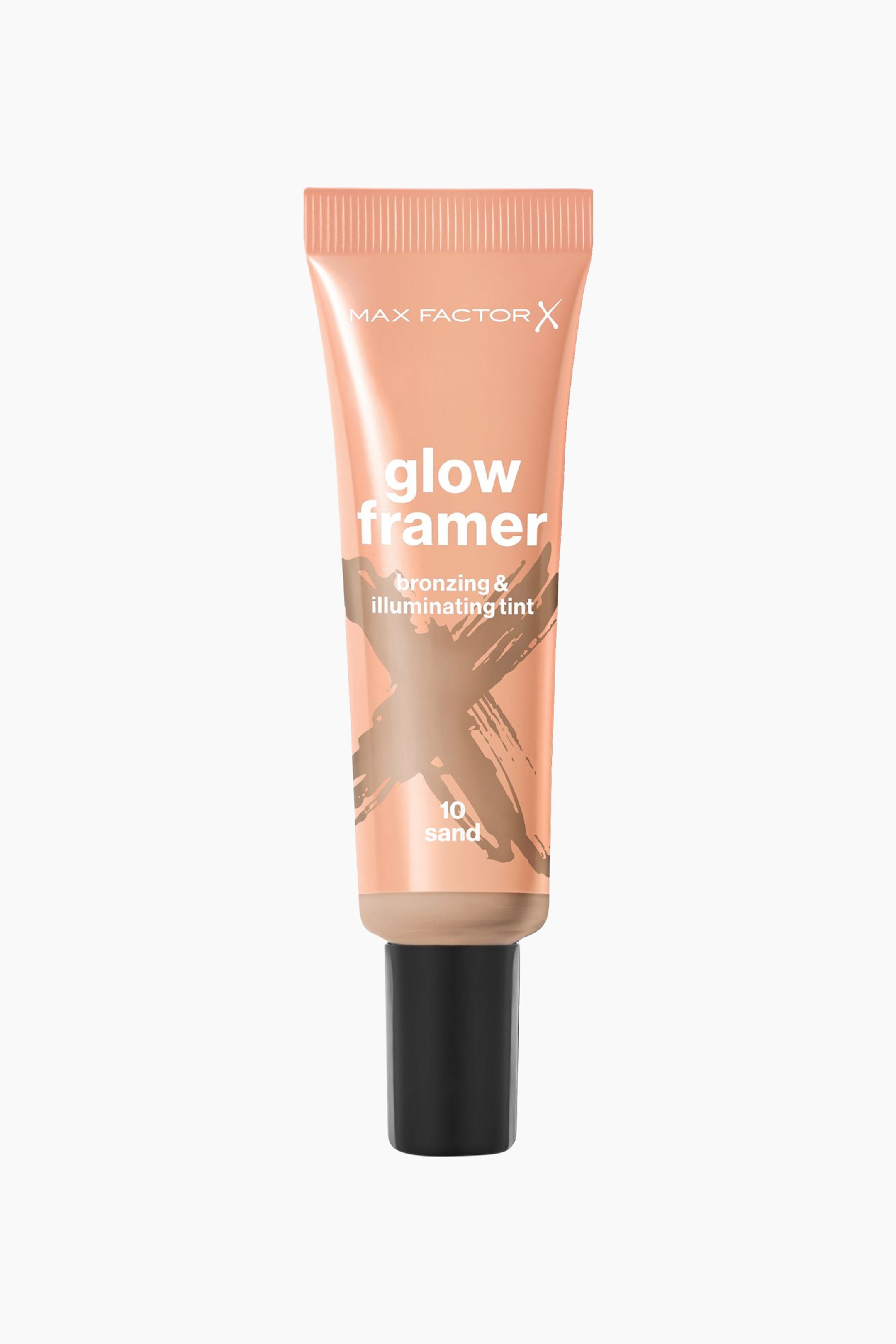 Se større billede: Et fersken-orange kosmetisk klemtube står oprejst, med fronten fremad. Tuben viser teksten "MAX FACTOR X glow framer bronzing & illuminating tint" og "10 sand". Et stort, lysebrunt X-formet penselstrøg-design dækker midten af tuben, og den har en sort cylindrisk hætte ved bunden.