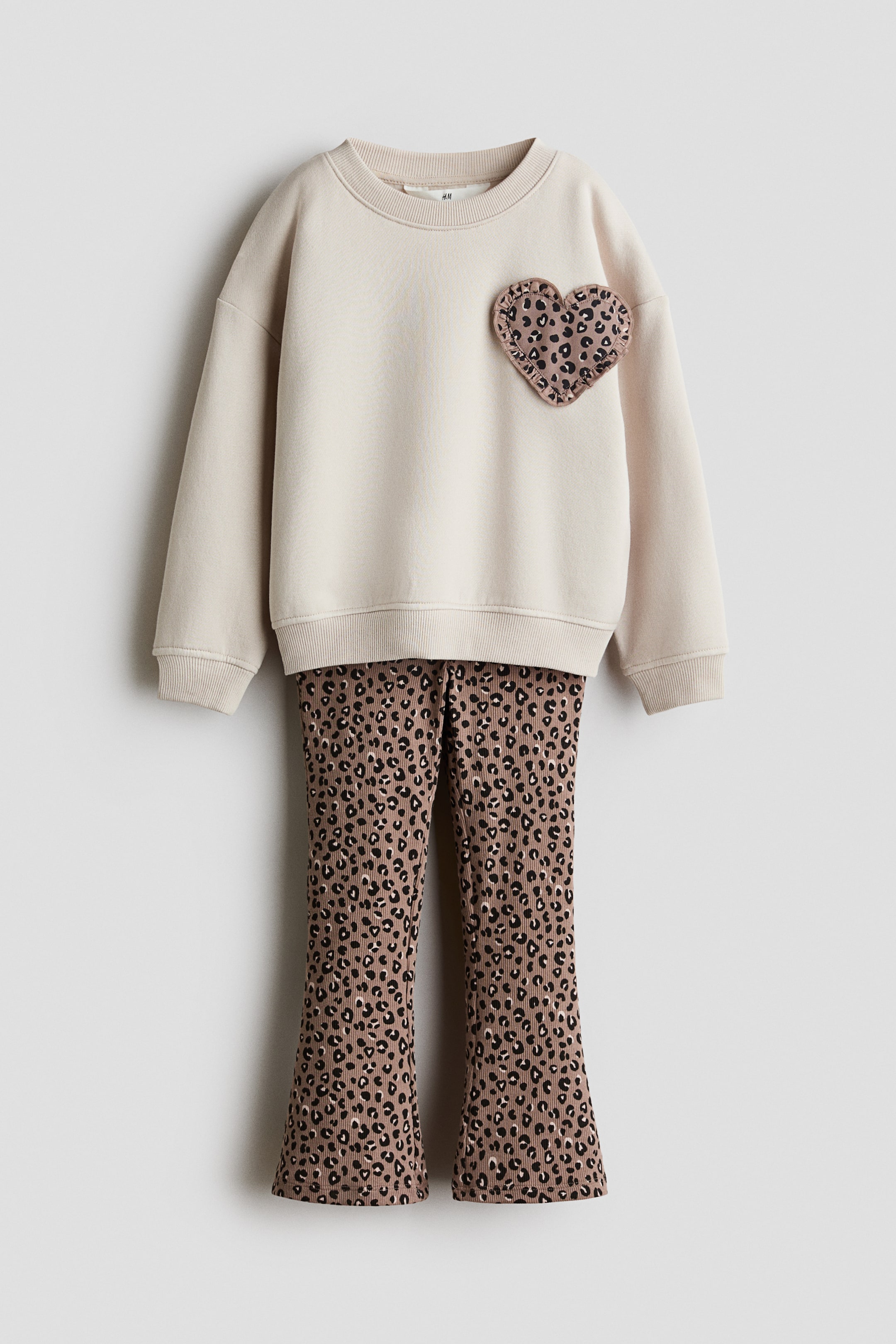 View larger image: A kids' two-piece set featuring a cream sweatshirt adorned with a leopard print heart patch and ruffled edge on the left chest, paired with matching ribbed pants showcasing a leopard print and a slight flare at the bottom.