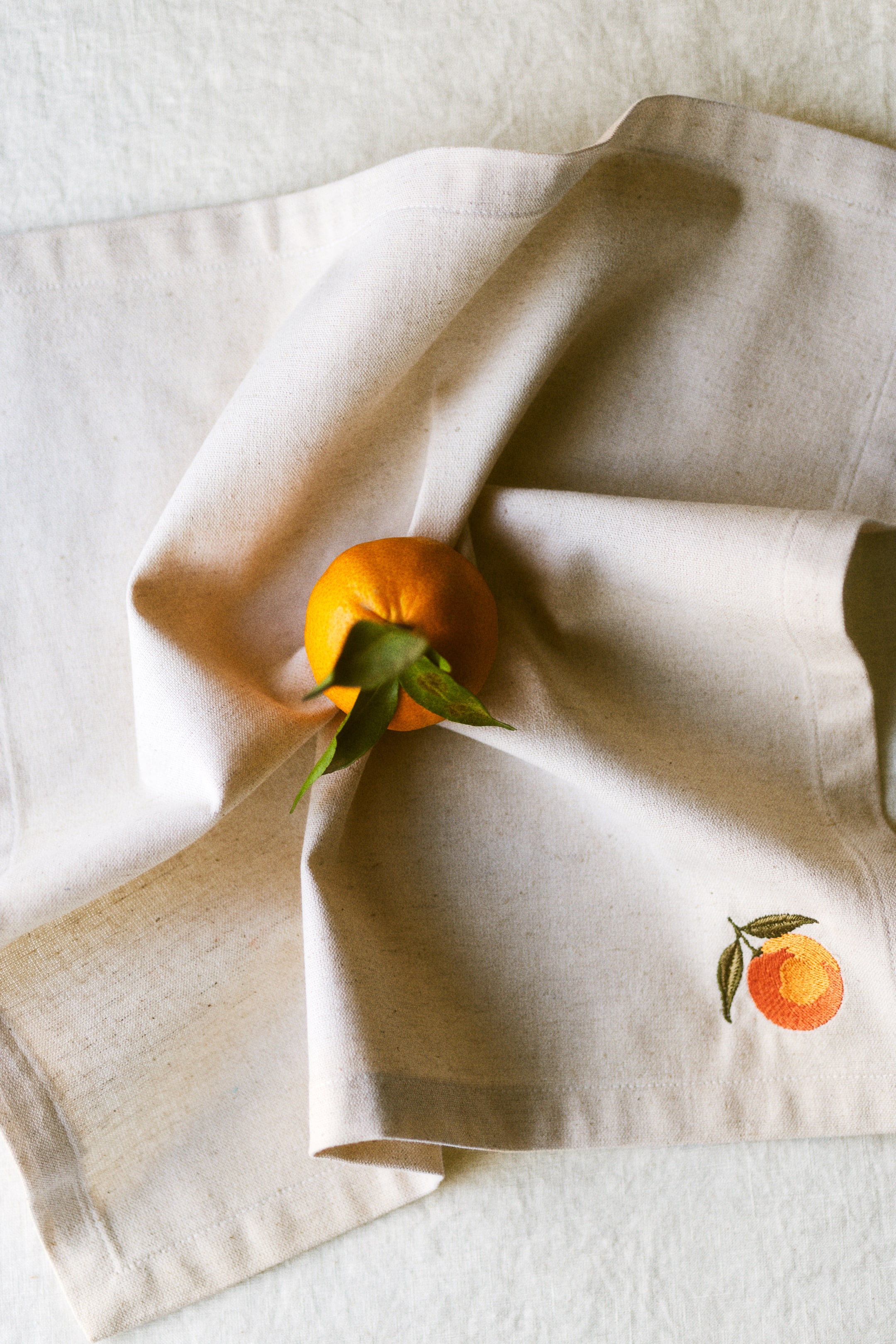 View larger image: A cream-coloured linen serviette is displaying soft folds, featuring an embroidered orange with green leaves in the bottom right. A fresh orange with leaves is resting on the serviette, creating a slight indentation.