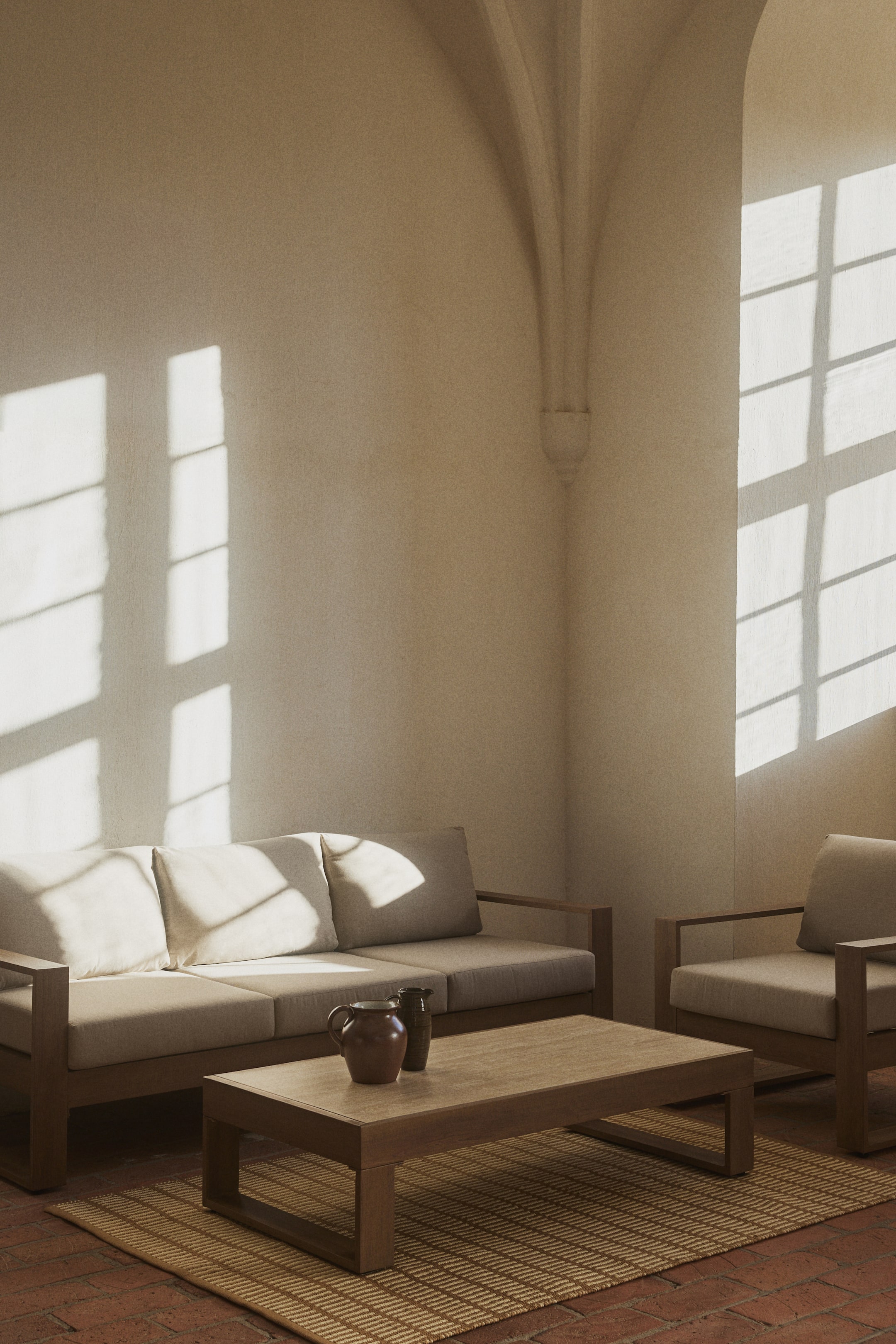 Se større billede: En tre-personers sofa og en matchende lænestol med lyse hynder på trærammer, arrangeret med et rektangulært træsofabord. To dekorative vaser hviler på sofabordet, som er placeret på et stribet tæppe.