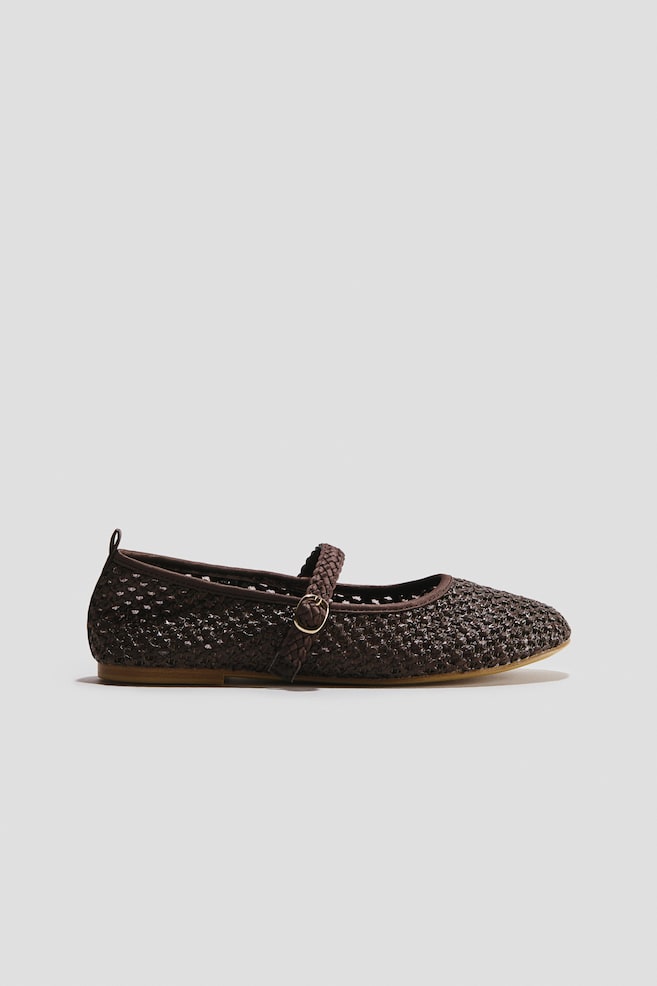 A single dark brown ballet flat in braided fabric is shown from a side profile view, featuring a rounded toe, a small heel pull loop, and a braided instep strap with an adjustable gold-toned metal buckle. The shoe rests on a thin, flat tan sole. - 1