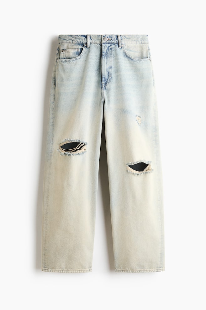 Ein Paar hell gewaschene Denim-Jeans mit weitem Bein hängt flach in frontaler Ansicht und zeigt eine weite Silhouette, eine reguläre Taille und das klassische Five-Pocket-Design. Die Jeans weist markante Used-Look-Risse mit ausgefransten Kanten an beiden Knien sowie eine dezente gelblich getönte Waschung im gesamten Stoff auf. - 1