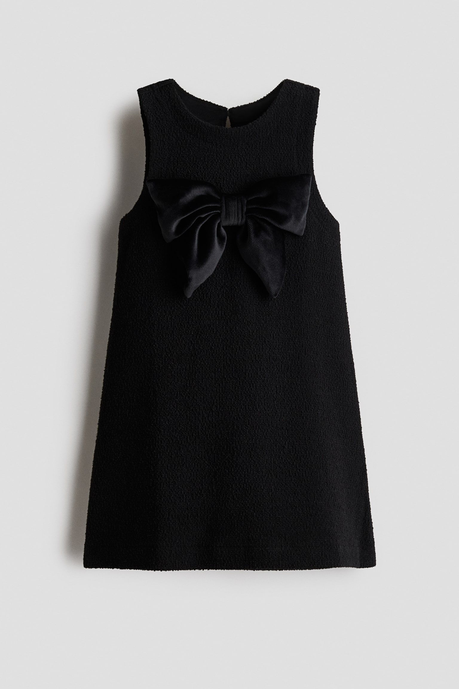 A black, sleeveless A-line dress in textured jersey is shown from the front, featuring a round neckline and a large decorative satin bow on the chest. A keyhole opening with a button-and-loop fastening is visible at the back of the neck.