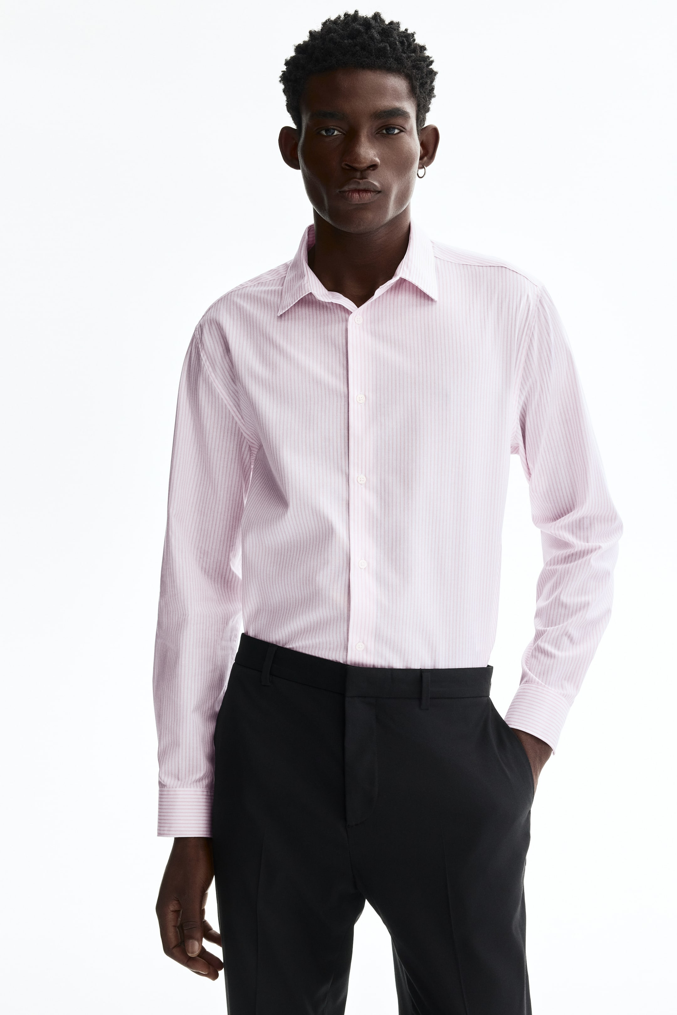 View larger image: The model is wearing a pink and white striped woven shirt, showcasing a turn-down collar, a classic button-front, and long sleeves with buttoned cuffs. The shirt is neatly tucked into black trousers, creating a tailored silhouette.