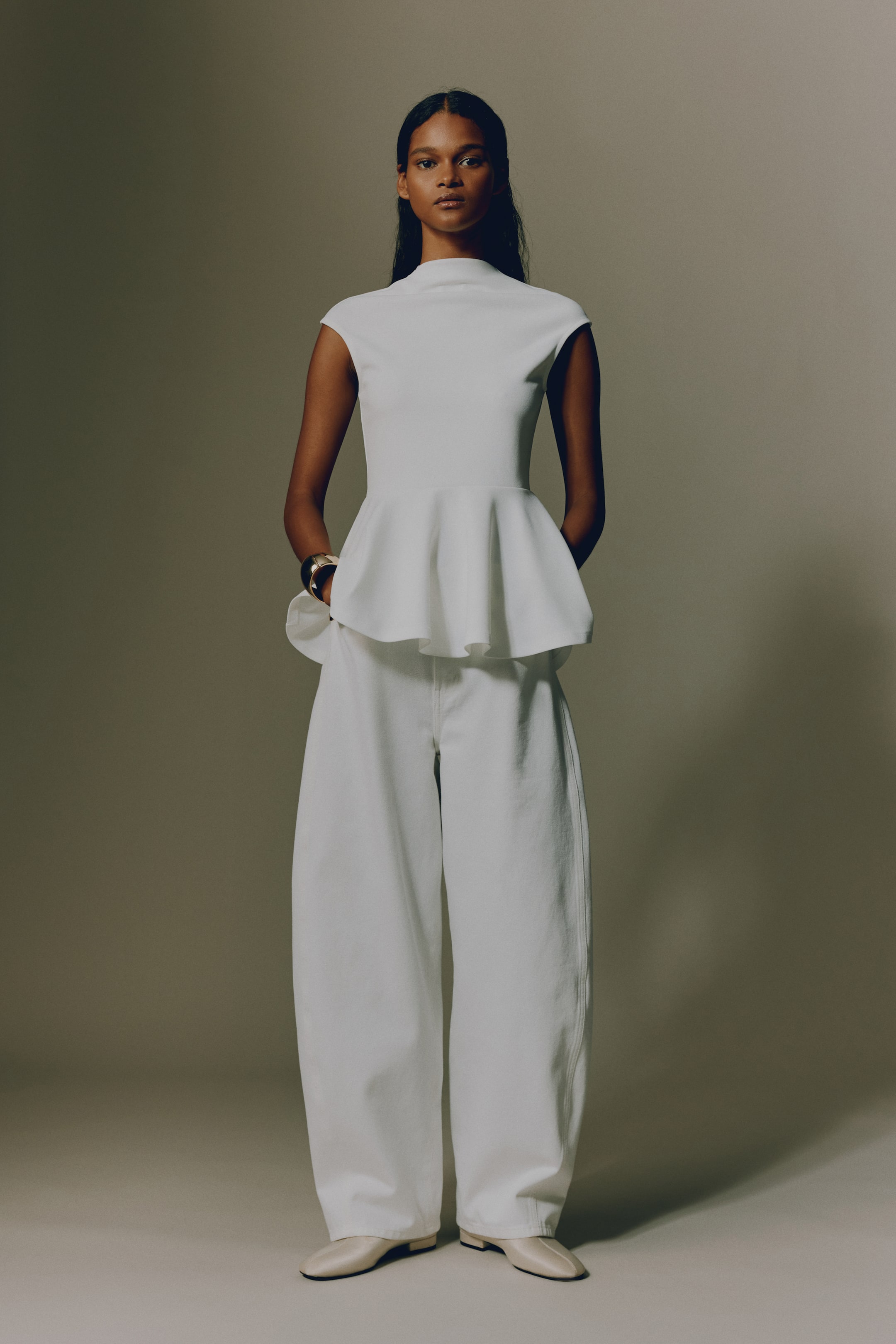 View larger image: A white cap-sleeve top featuring a mock neck and a flared peplum hem, paired with white wide-leg pants. The pants have a relaxed, full-length fit and fall over light cream flat shoes.