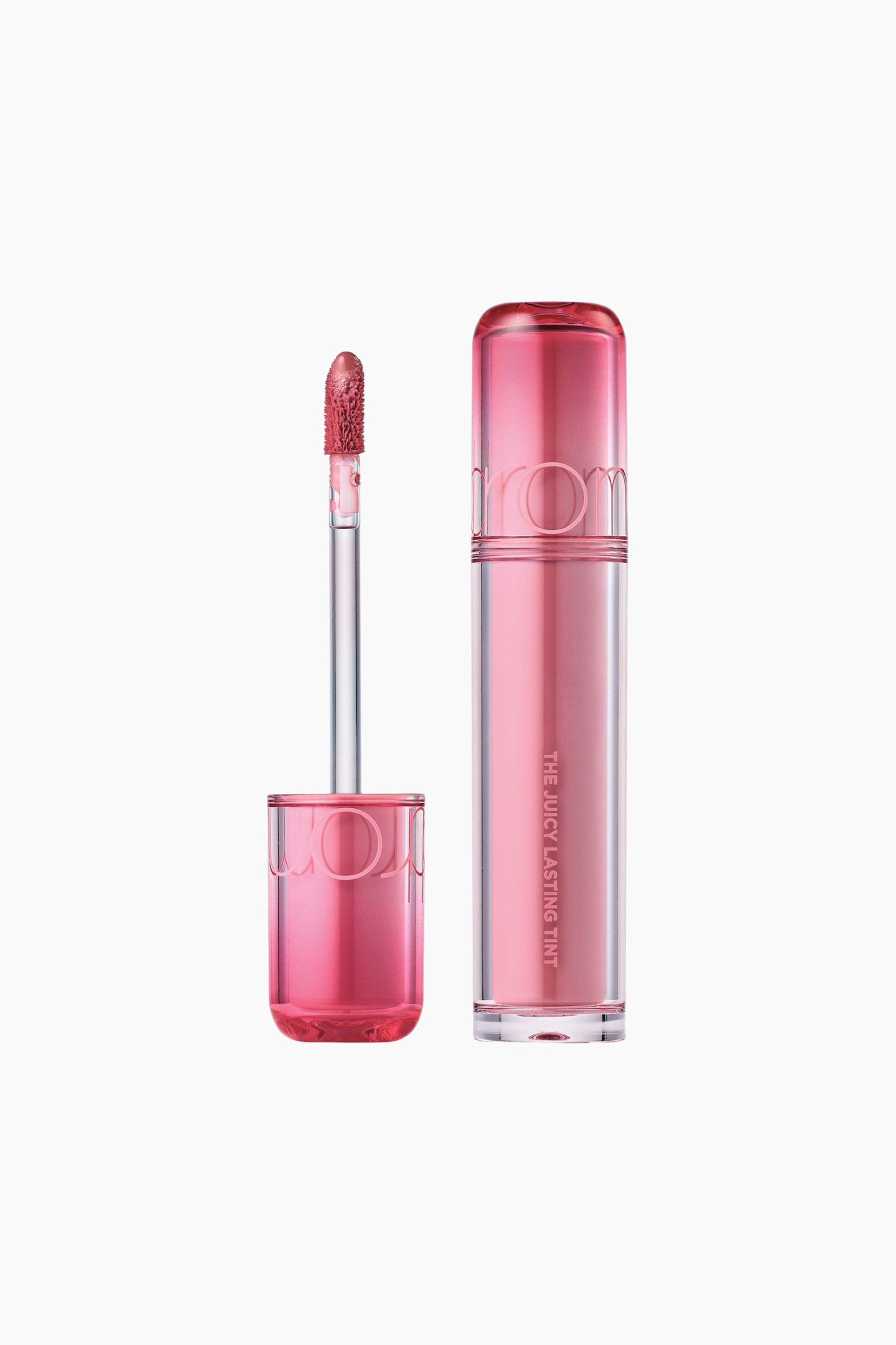 Vis større bilde: En sylindrisk rom&nd The Juicy Lasting Tint står oppreist og har et klart hylster fylt med en lys rosa tint. Den gjennomsiktige rosa hetten ligger ved siden av hylsteret og holder den teksturerte doe-foot-applikatoren, som er dekket av den samme rosa tinten.