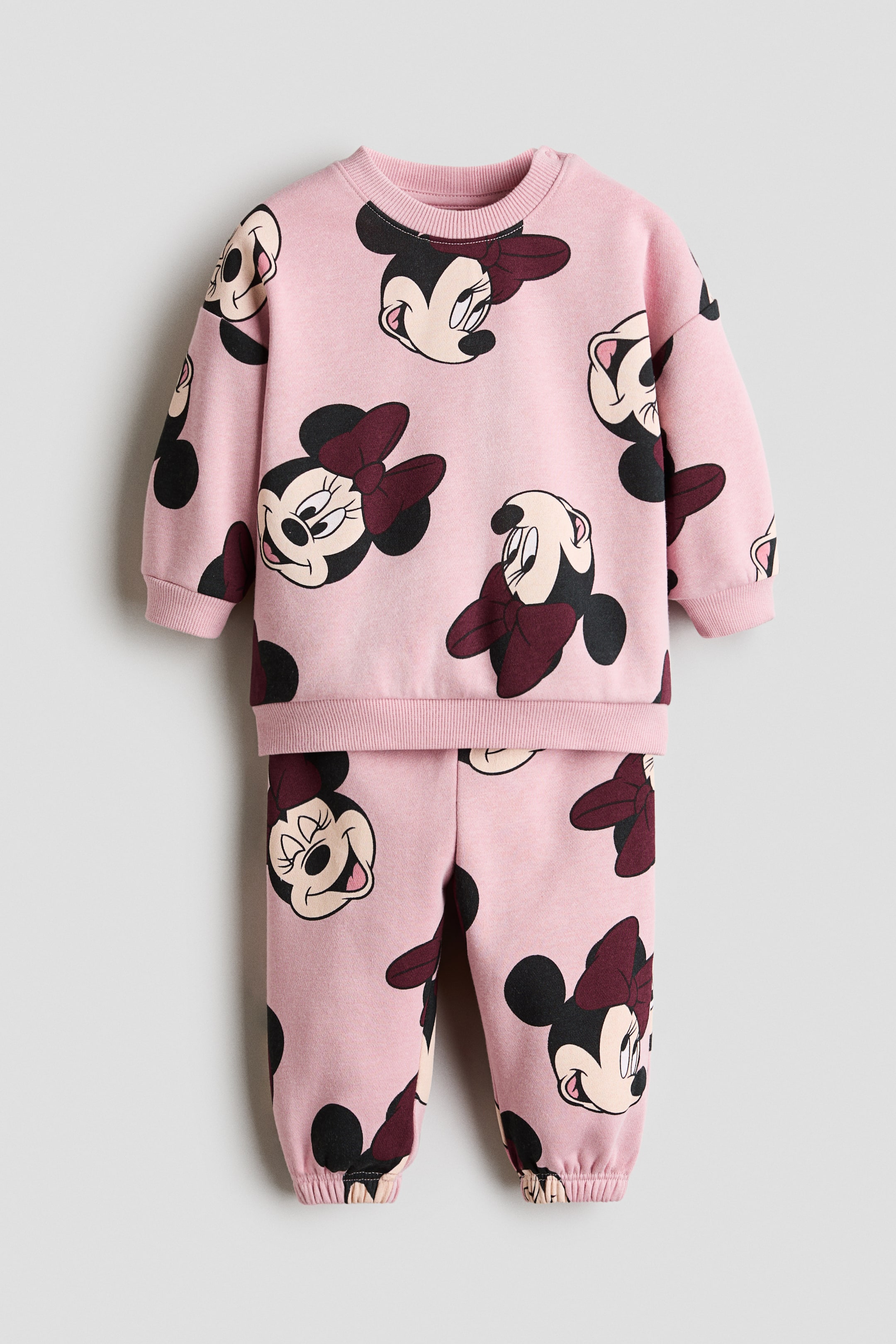 View larger image: This 2-piece pink printed sweatshirt set is showcased laid flat, facing forward. The set boasts a repeating pattern of Minnie Mouse heads adorned with maroon bows. The sweatshirt features a ribbed crew neck, ribbed cuffs, and hem, while the matching pants are designed with elasticated ankle cuffs.
