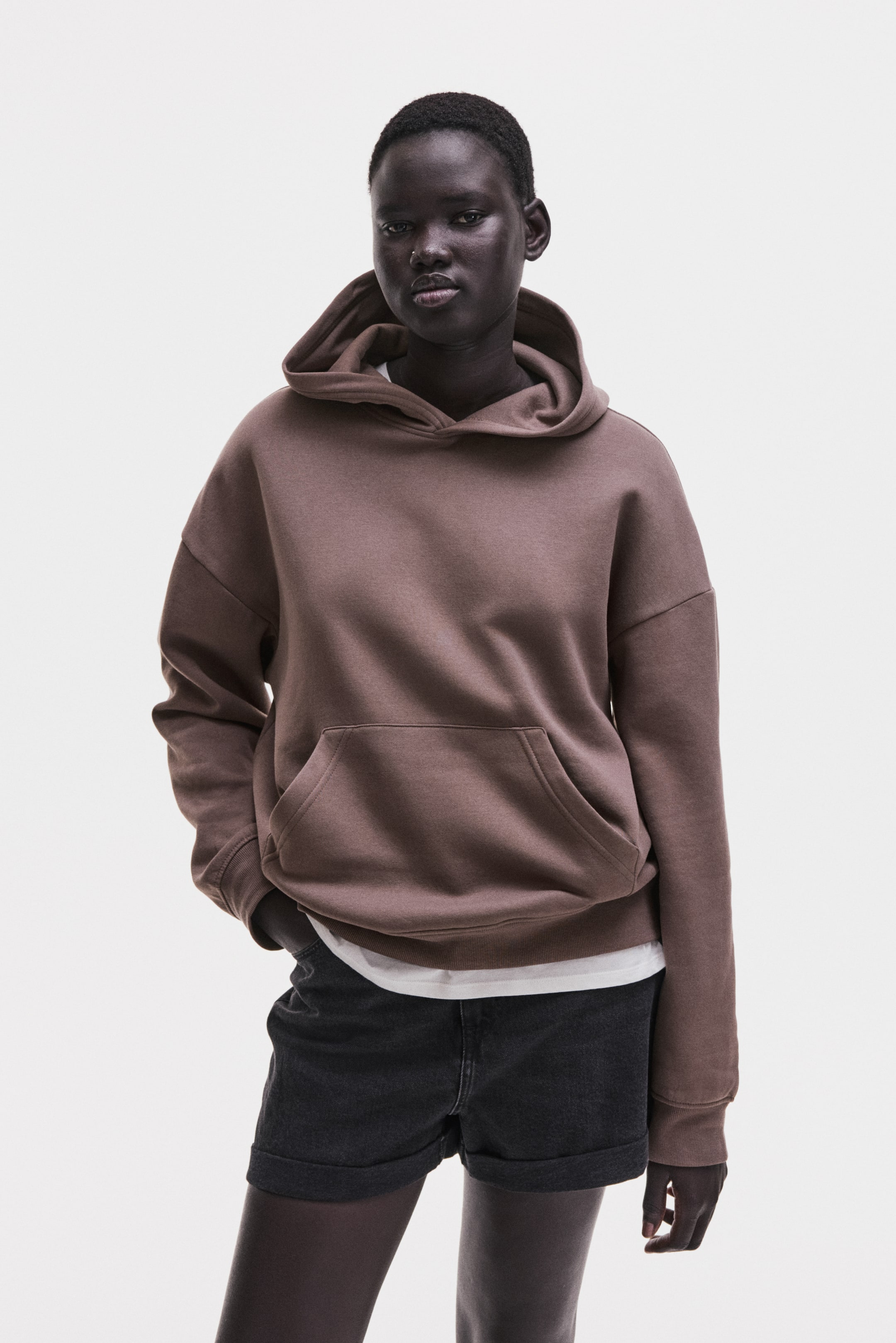 View larger image: The brown oversized hoodie is shown from the front, featuring a large kangaroo pocket, ribbed cuffs, and a ribbed hem. A white T-shirt is layered underneath, visible at the neckline and just below the hoodie's hem.