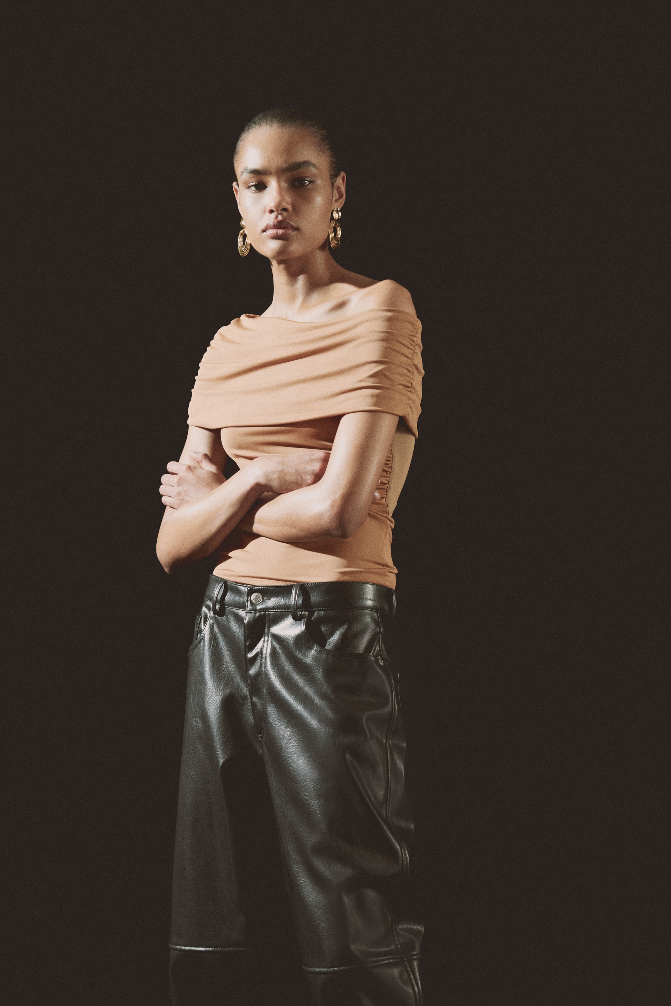View larger image: The beige off-the-shoulder top showcases a draped neckline across the chest and ruching on the right shoulder. The black high-waisted trousers, crafted in a leather-look material, feature belt loops, front pockets, and seam detailing on the knees.