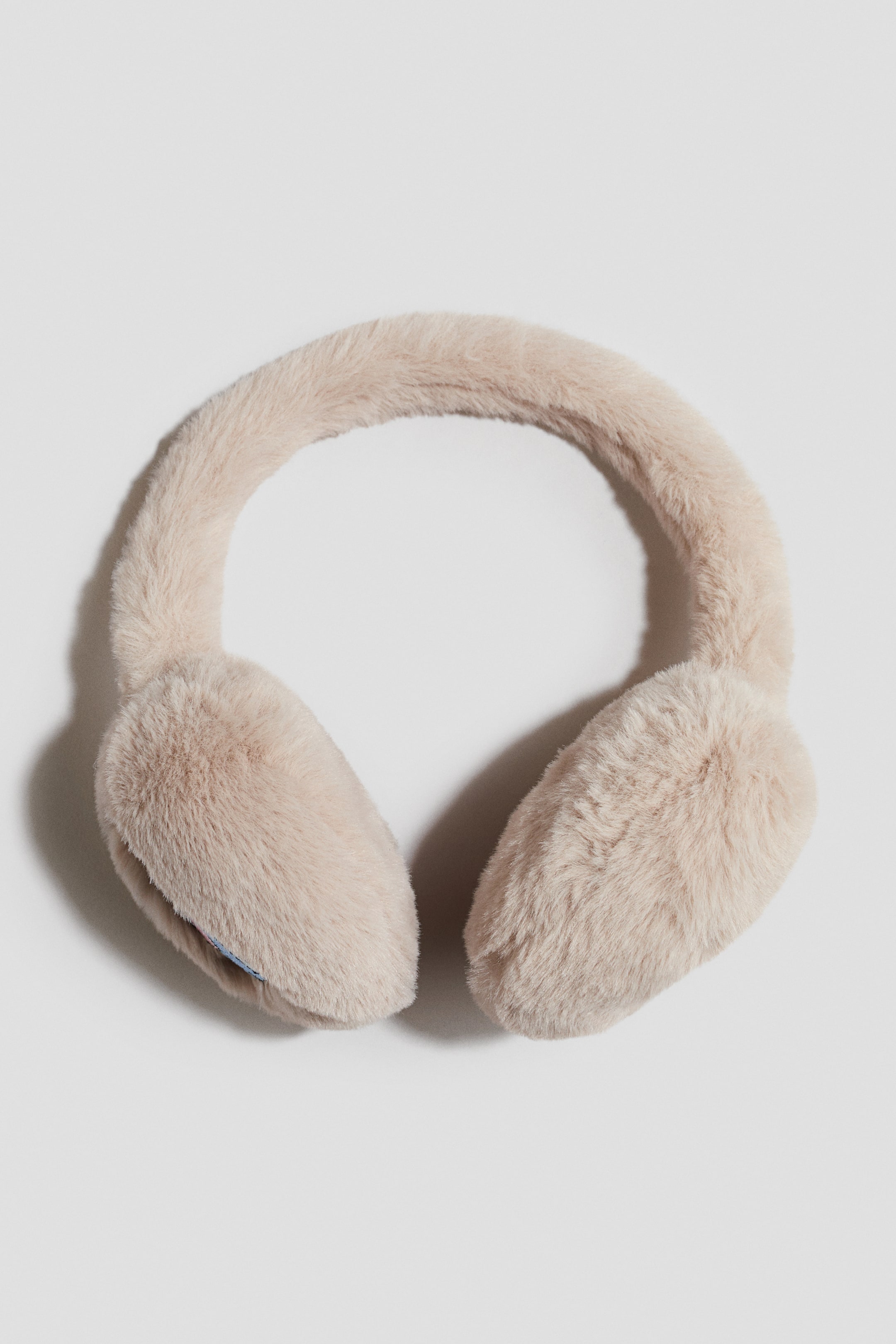Voir une image plus grande: Des cache-oreilles duveteux de couleur beige pâle sont présentés de face, avec un bandeau recouvert de fausse fourrure moelleuse reliant deux oreillettes arrondies et généreusement rembourrées.