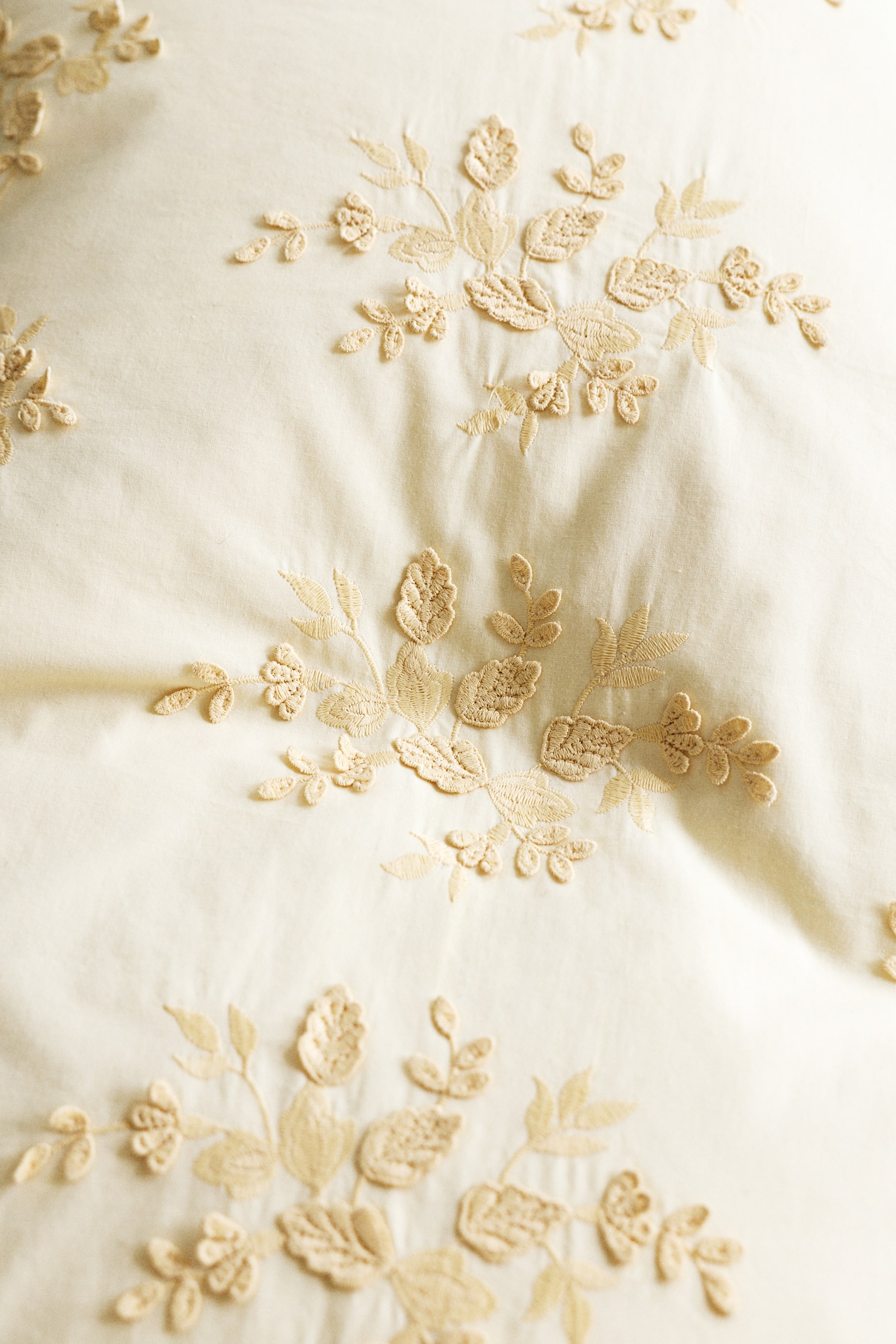 Visa större bild: Ett örngott visar broderade blommönster och bladmotiv över sitt mjuka bomullstyg. Broderiet framträder i beige tråd, vilket står i kontrast till det ljusa krämfärgade tyget.