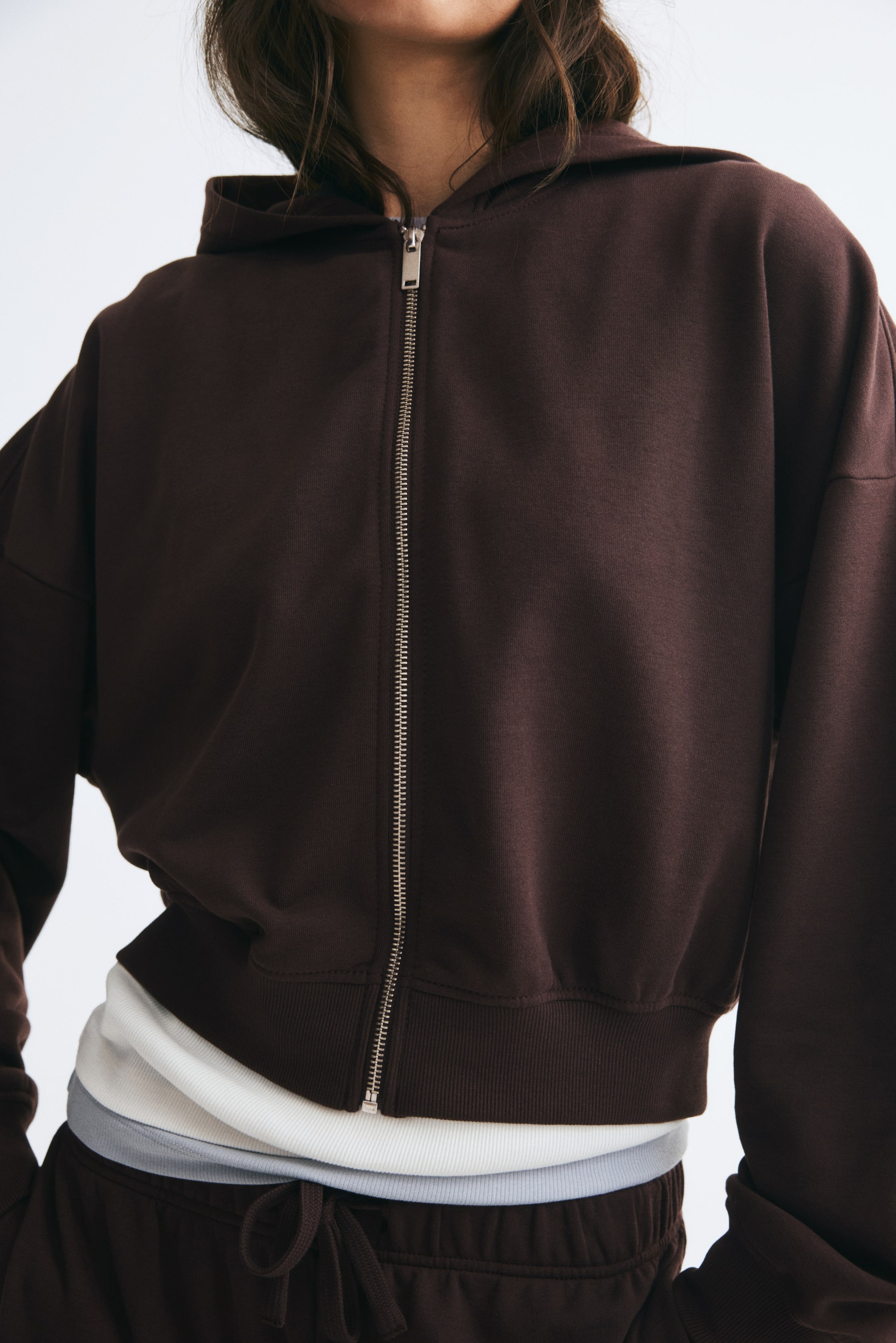 View larger image: The model is wearing a short, dark brown zip-up hoodie featuring low dropped shoulders, a silver front zipper, and a ribbed hem. The hoodie is layered over a white ribbed top and a light grey underlayer, and paired with matching brown sweatpants.